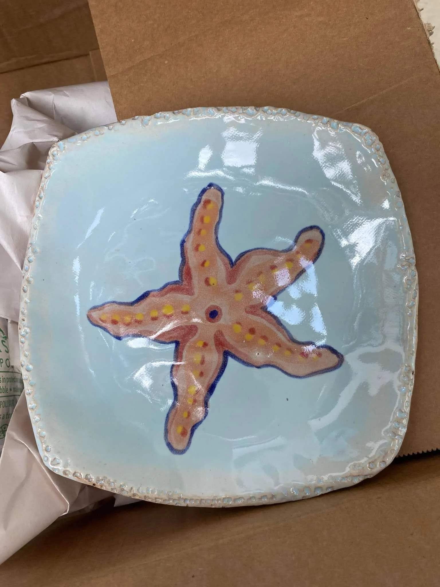 Square ceramic plate with a starfish illustration in the center, featuring five arms, yellow dots, and a dark blue outline, on a light blue background.