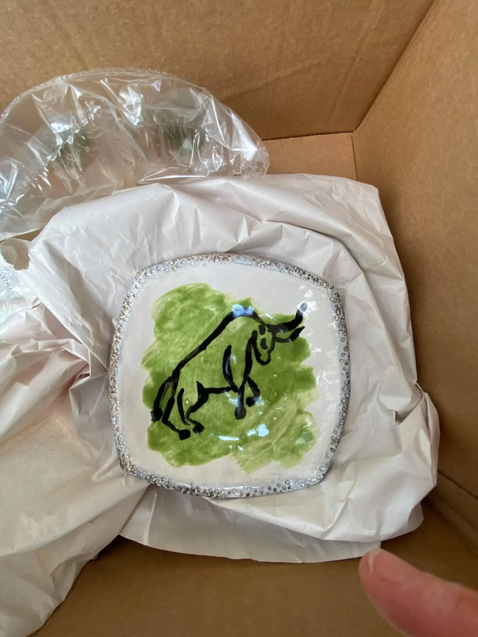 Decorative ceramic plate with a hand-painted image of a black elephant standing on green grass, inside a cardboard box lined with tissue paper.