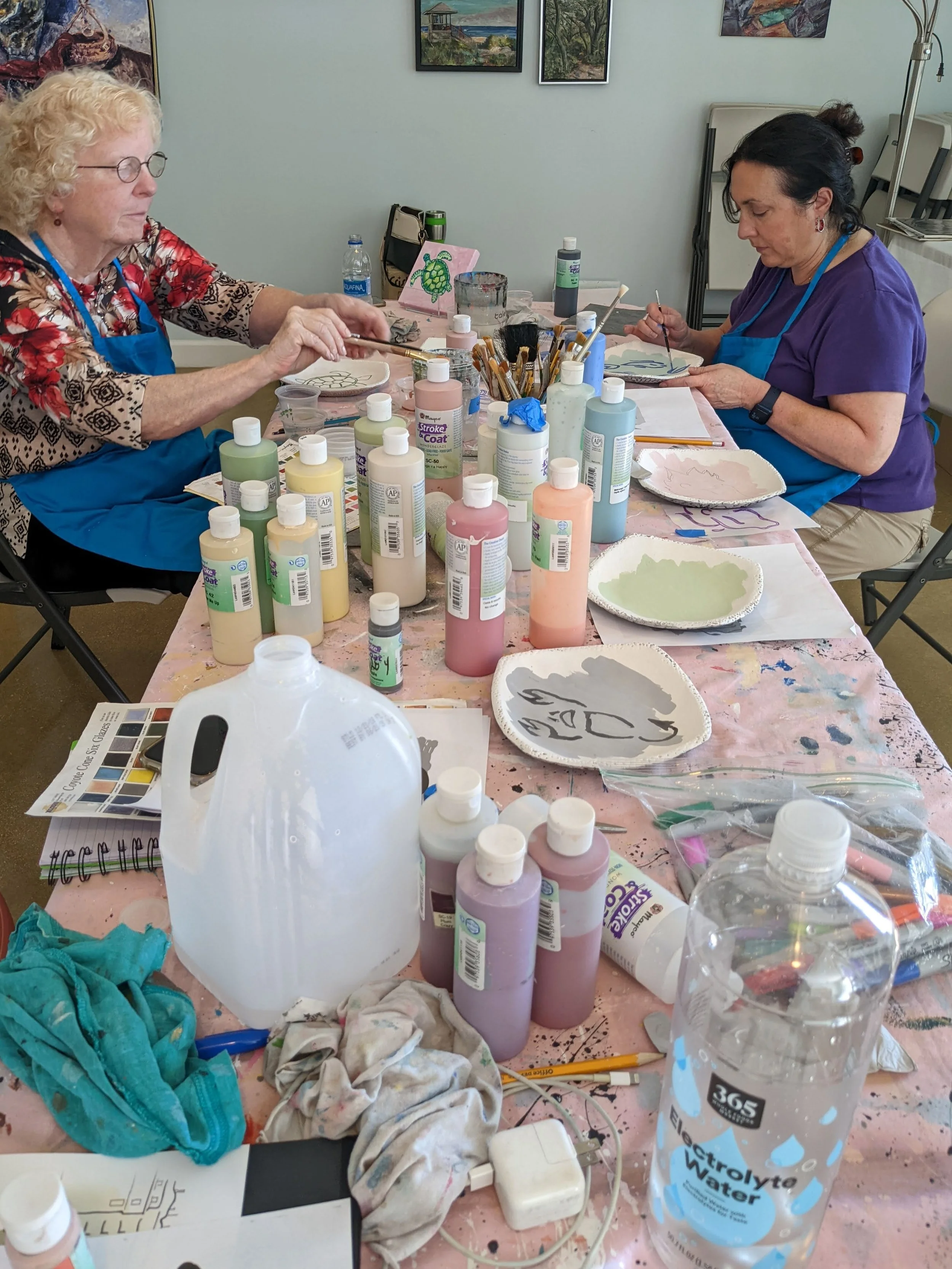 Two women painting ceramic plates at a table covered with art supplies, including bottles of paint, brushes, and water bottles, in an art studio or classroom.