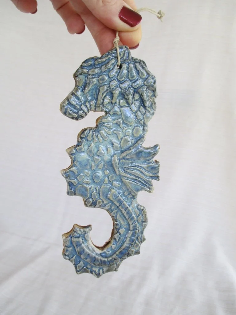 A ceramic seahorse ornament painted in blue shades, held by a person's hand with red nail polish, against a plain background.