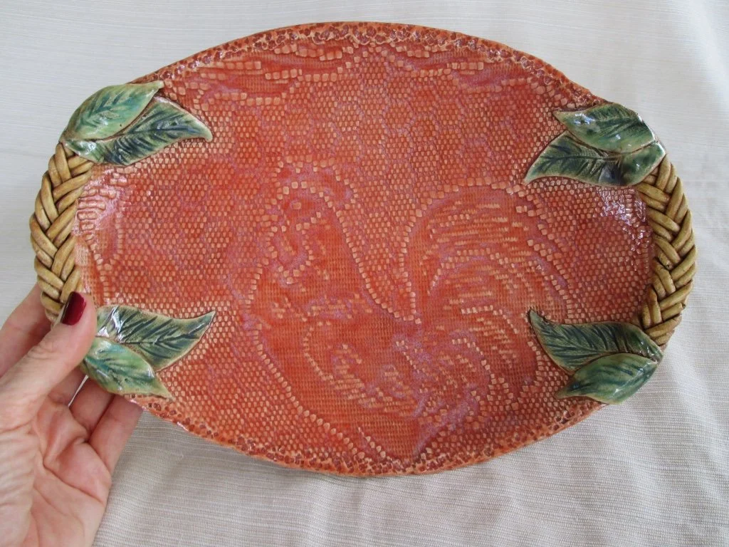 Decorative oval ceramic platter with a rooster design in the center, embellished with four green ceramic leaves at each corner, and a braided woven rim.