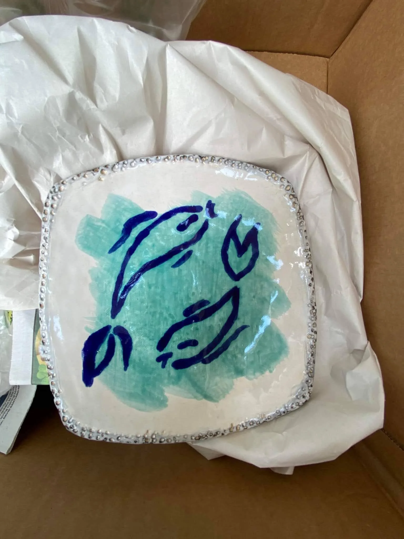 Decorative square cake with a white base, blue handwritten words, green watercolor background, and a border of silver edible beads, wrapped in white tissue paper inside a cardboard box.