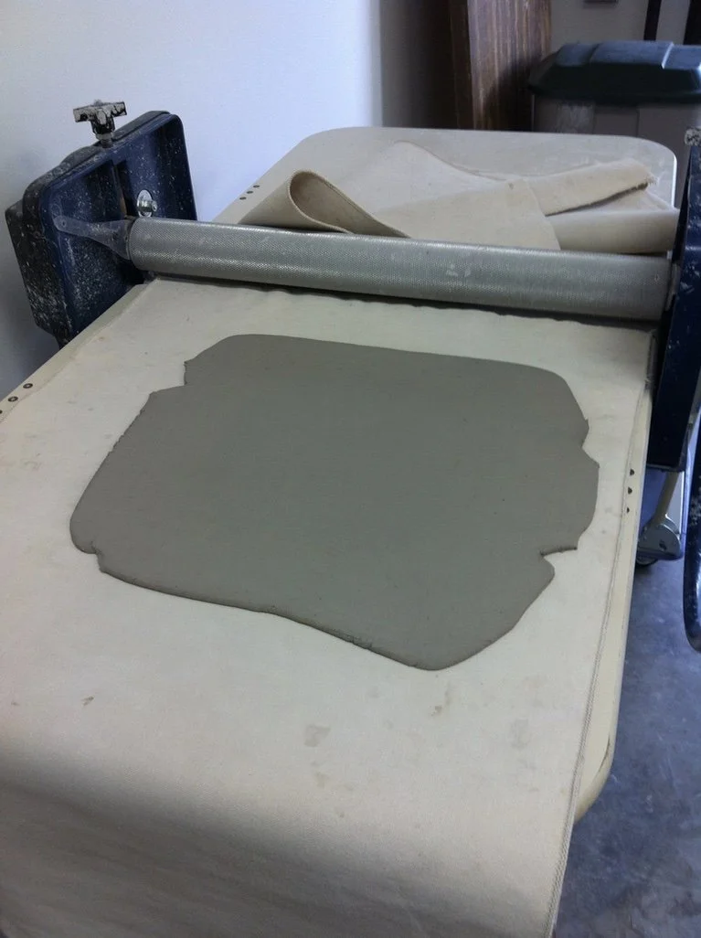 A table with a flattening machine, patches of clay or dough, and a rolling pin.
