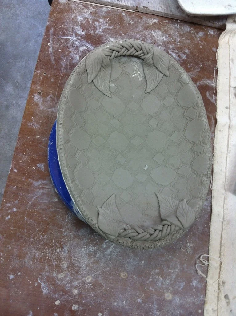 A ceramic oval plaque with embossed leaves and chain design, sitting on a worktable.