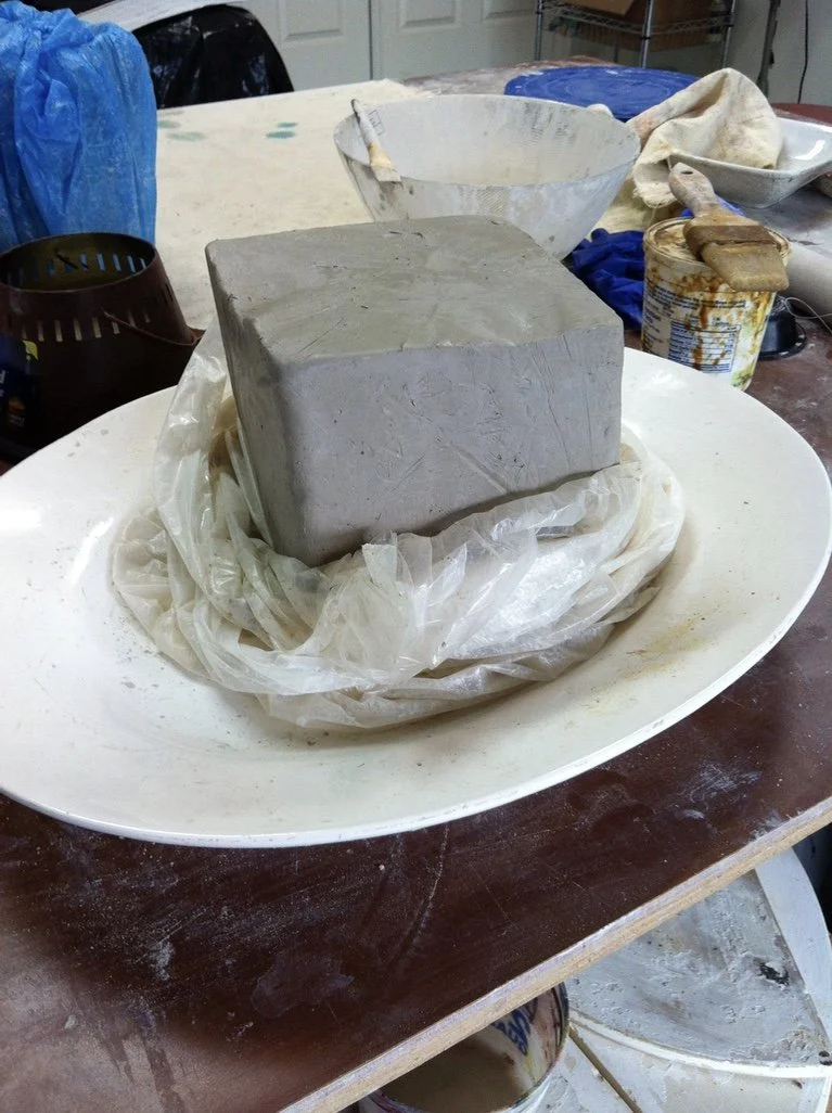 A block of clay on a plastic sheet on a white tray, surrounded by pottery tools and bowls on a workbench.