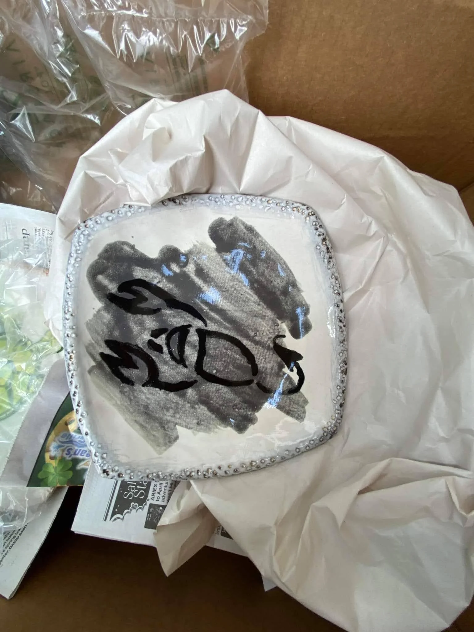 Decorative plate with a black and white handprint design, surrounded by a bead-like border, placed on tissue paper inside a box.