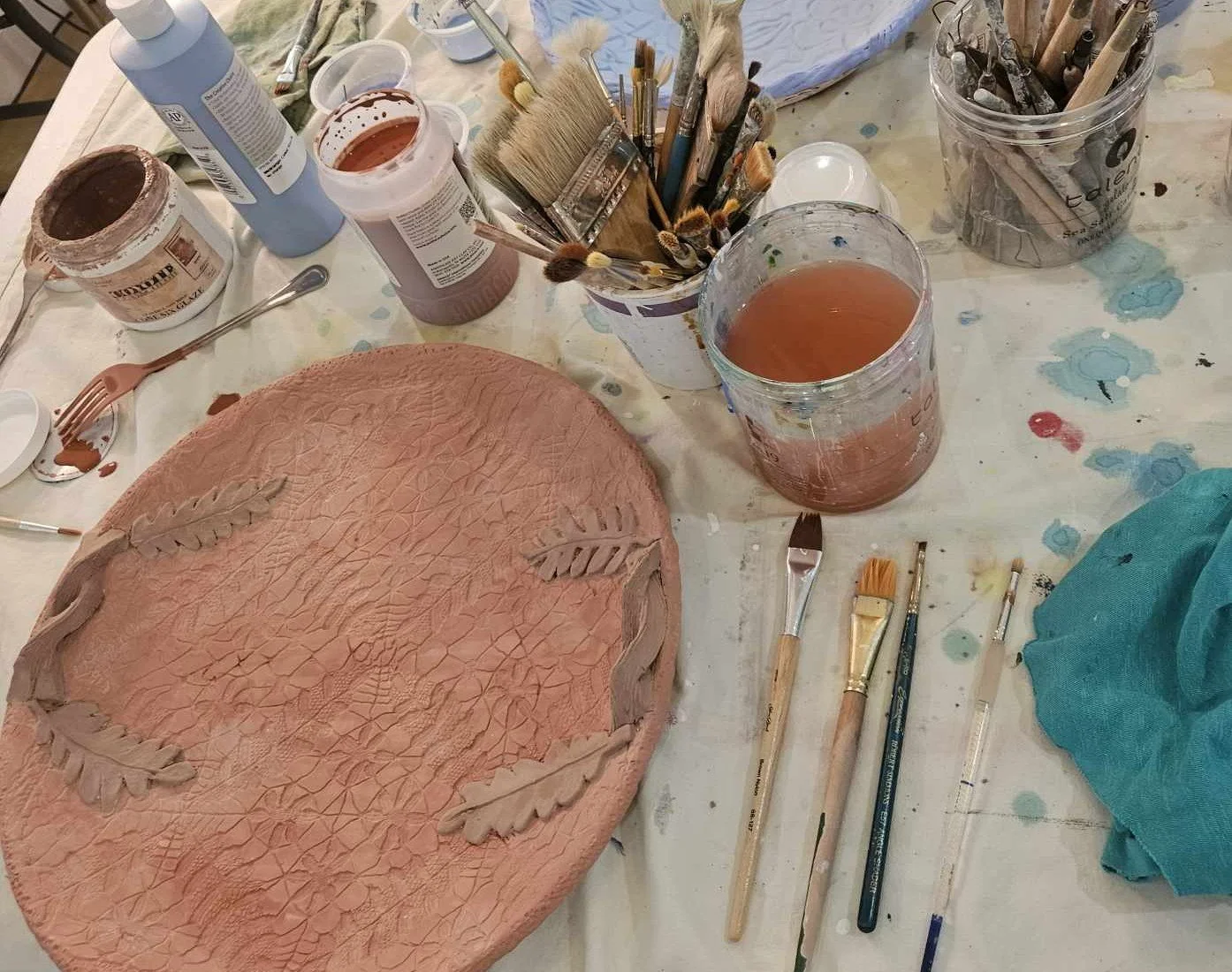 A workspace with clay art project, brushes, and jars of watercolor paints and glazes on a table covered with paint stains.