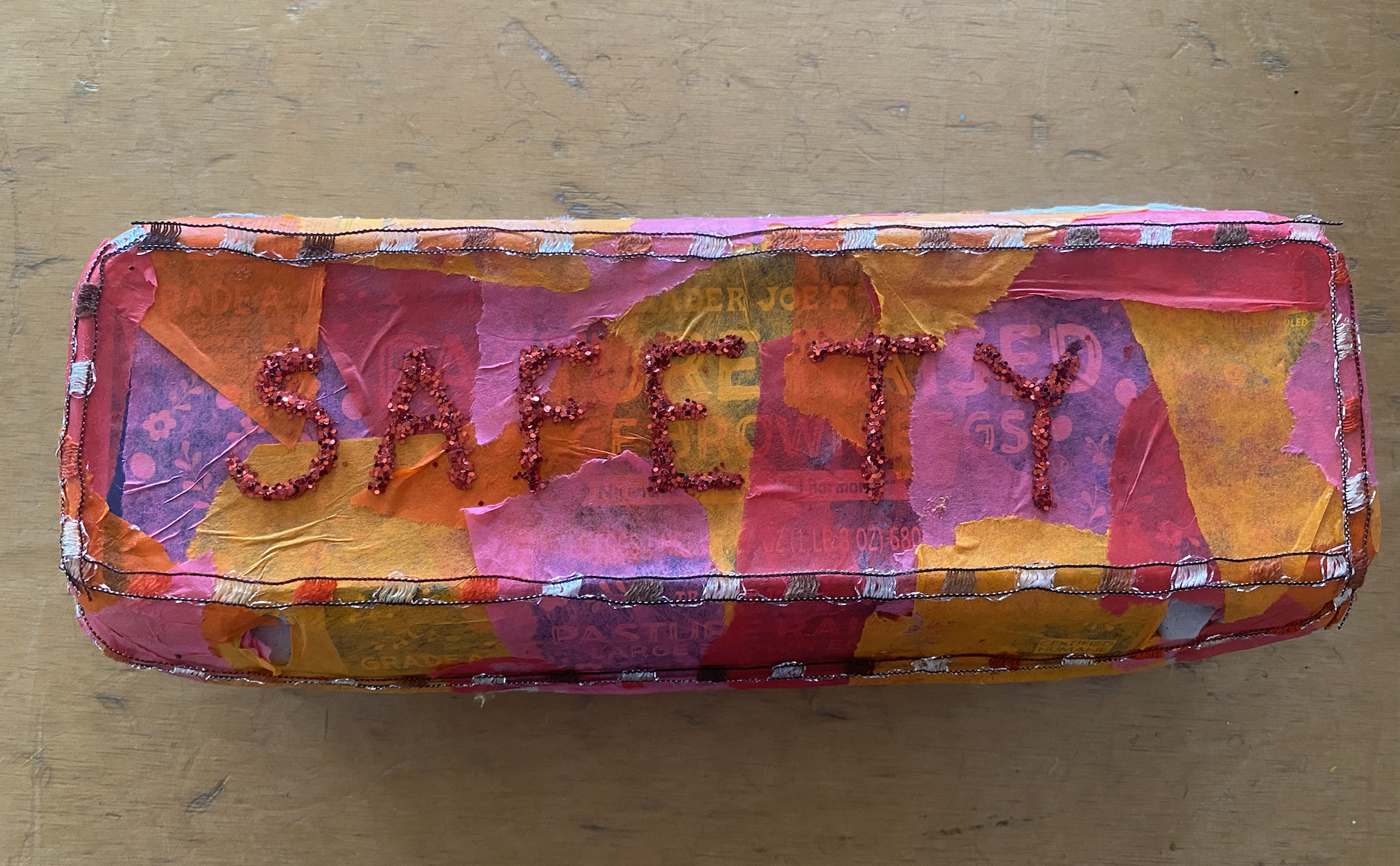 A rectangular object covered with torn colorful paper and decorated with pink beads that spell out the word "SAFETY."