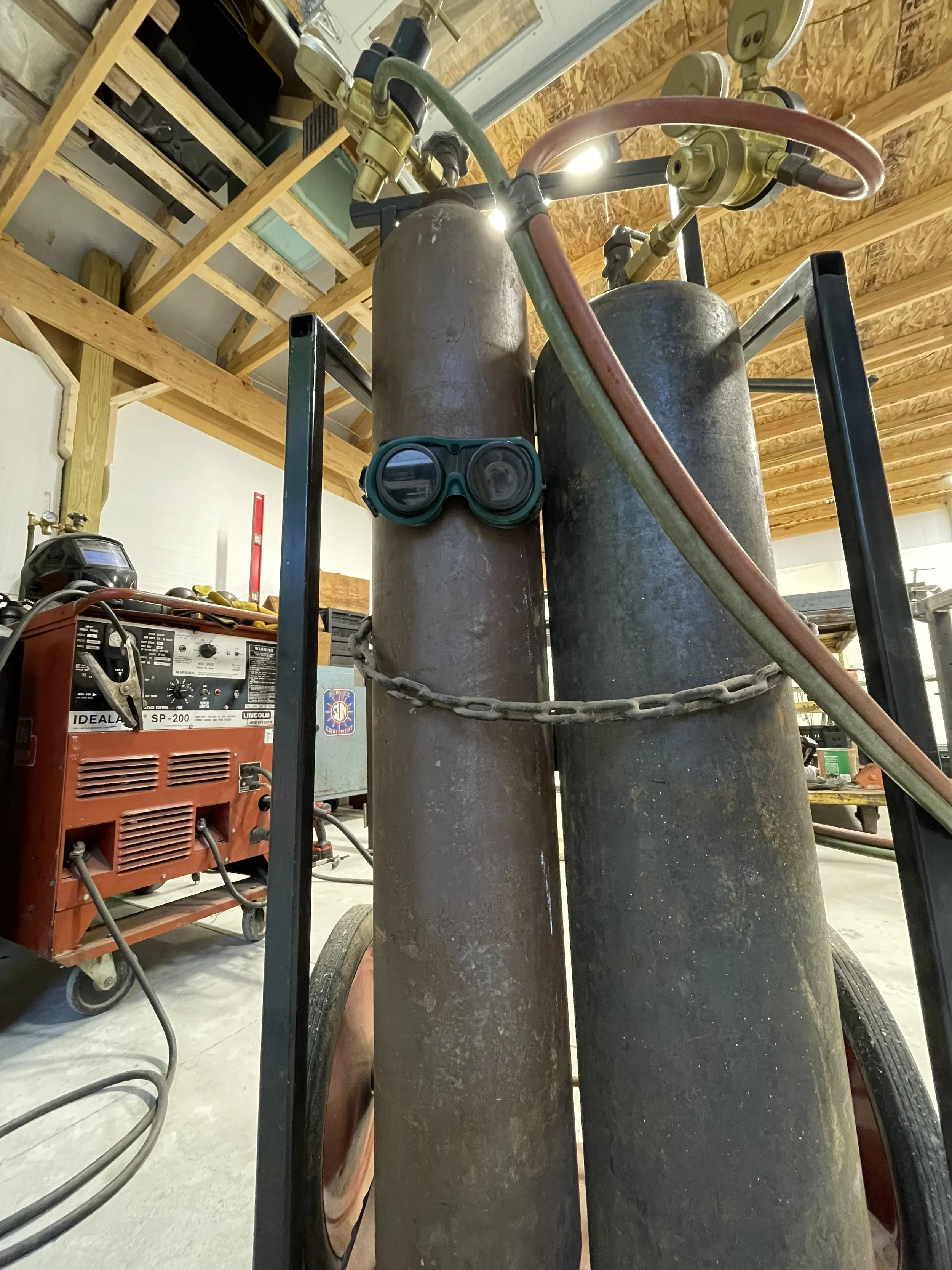 Two large, rusted oxygen or acetylene tanks mounted on a wheeled cart with welding goggles hanging on one of the tanks, in a workshop with wooden ceiling beams and various tools and equipment in the background.