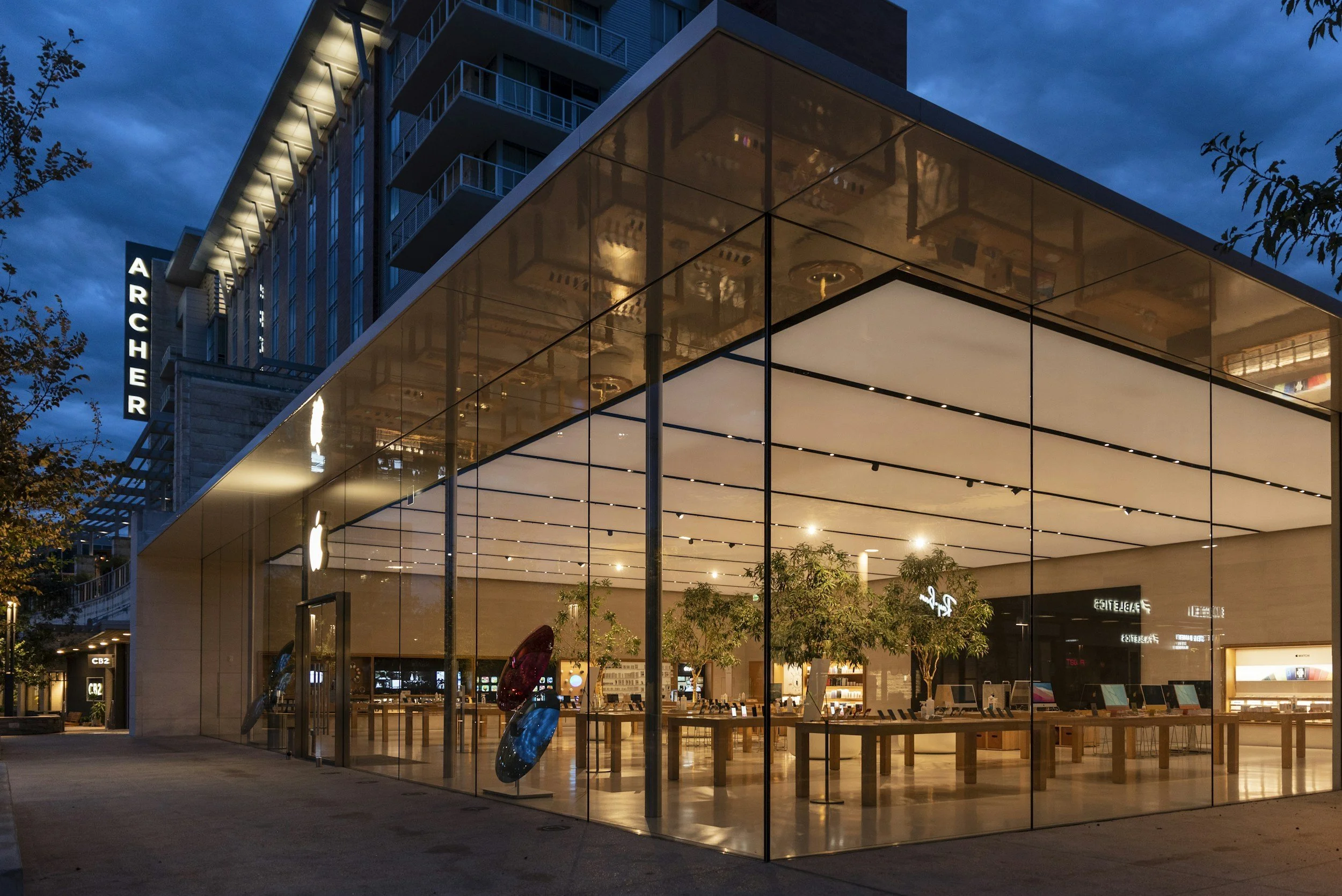 Apple Store with a glass facade, showing tables of iPads and trees inside at night, with a blue sky and nearby buildings, including an Archer sign.