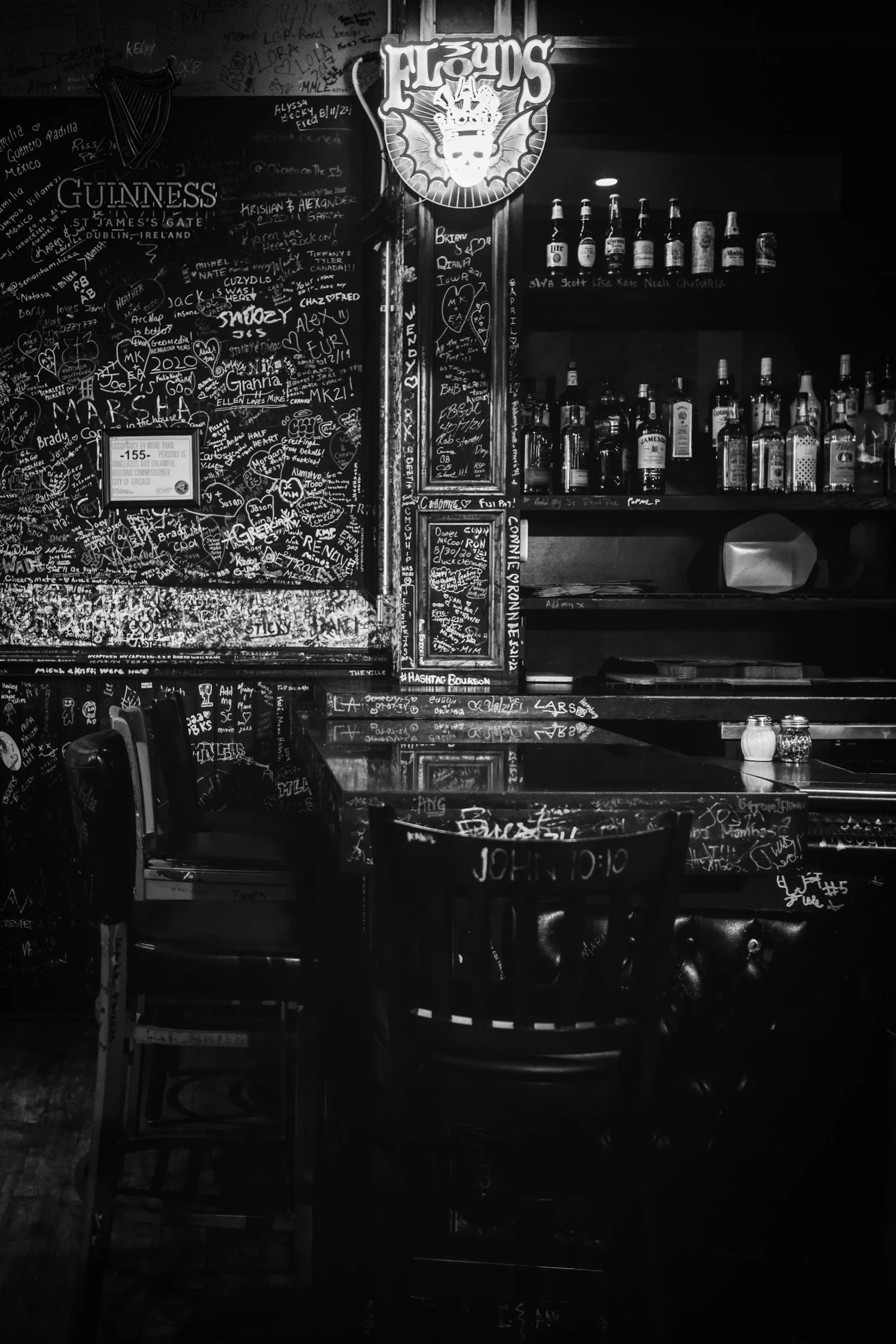 A black and white photo of a bar interior with a chalkboard wall filled with handwritten notes, signatures, and doodles. There are shelves stocked with liquor bottles, and a neon sign of a creature with the word 'ALOUUDS' above it. The bar counter has a few chairs, and salt and pepper shakers are on the counter.