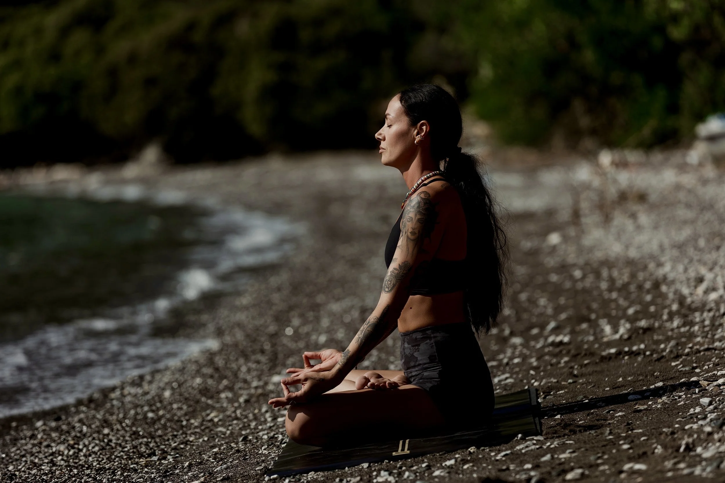 A woman with tattoos practicing meditation on a yoga mat on a rocky beach near the water, with trees in the background