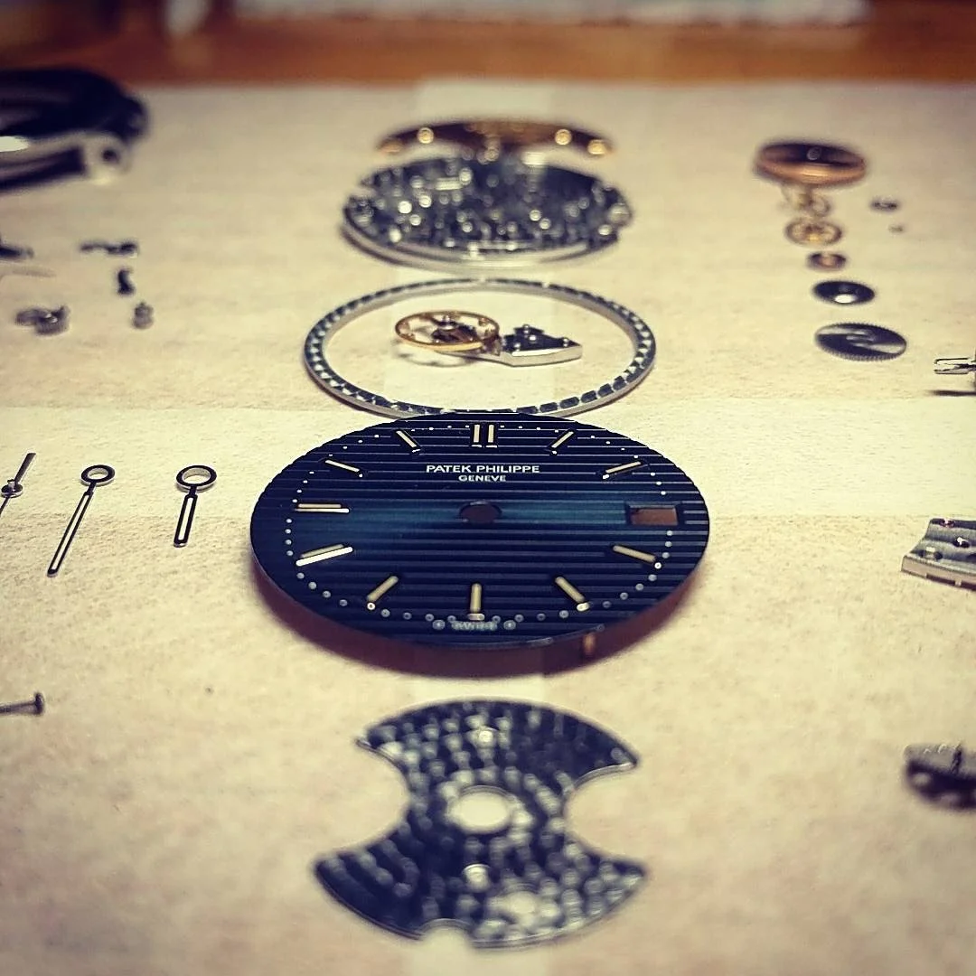 Disassembled parts of a Patek Philippe wristwatch laid out on a light-colored surface, including the watch face, gears, and small components.