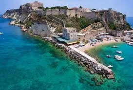 A coastal city with historic stone buildings, a small beach, and boats docked near the shore, with cliffs and ocean in the background.