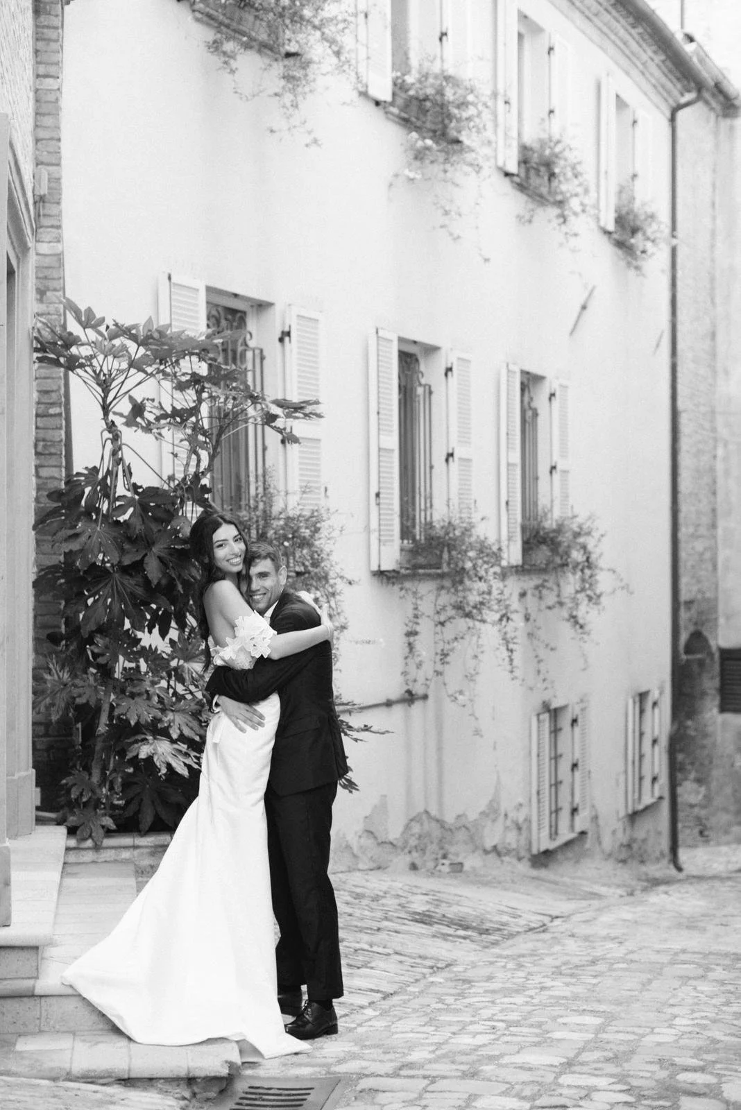 A black and white photo of a smiling woman in a wedding dress embracing a man in a suit on a cobblestone street with old buildings and shutters.