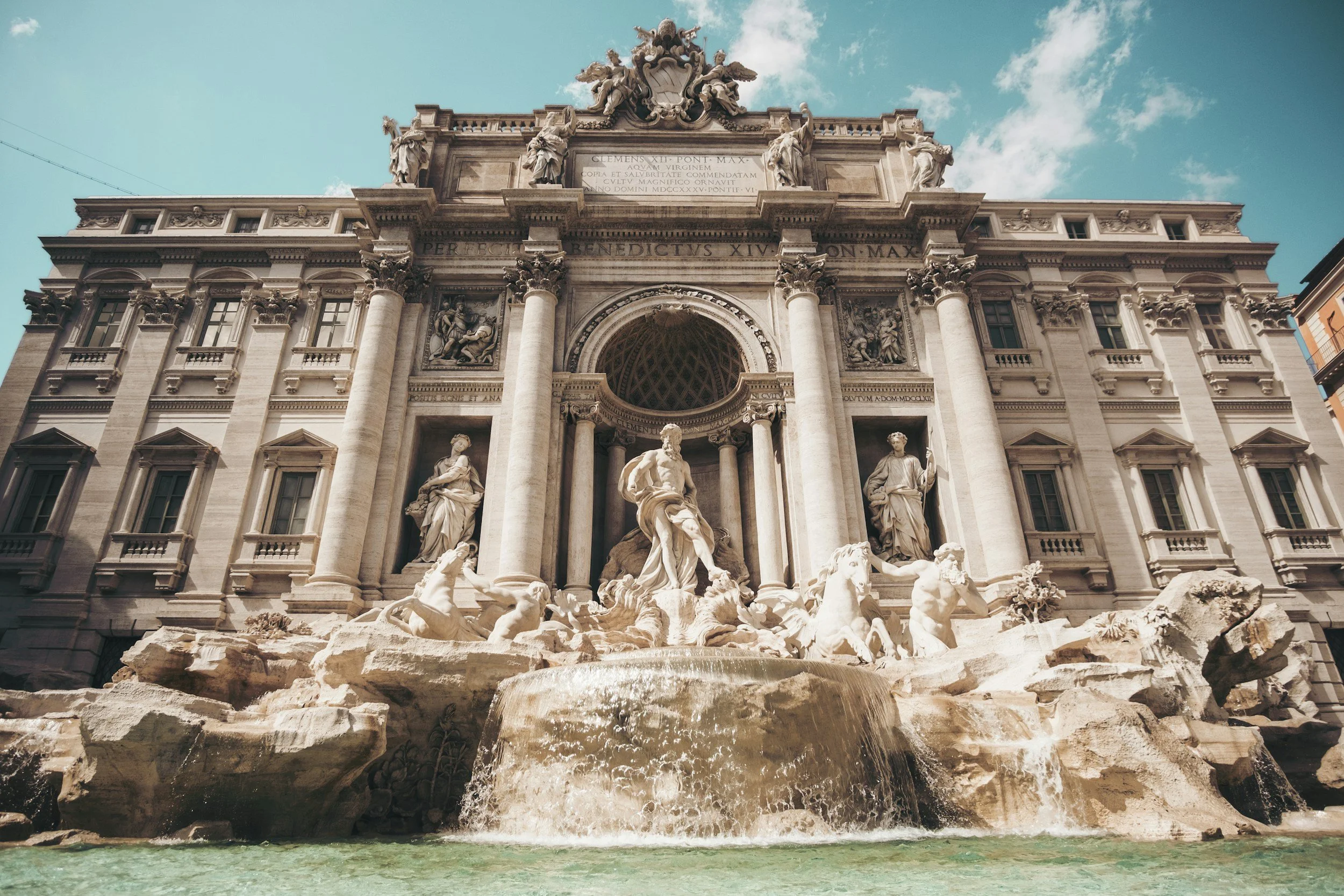 The Trevi Fountain in Rome, Italy, featuring intricate sculptures of mythological figures, cascading water, and an ornate facade against a partly cloudy sky.