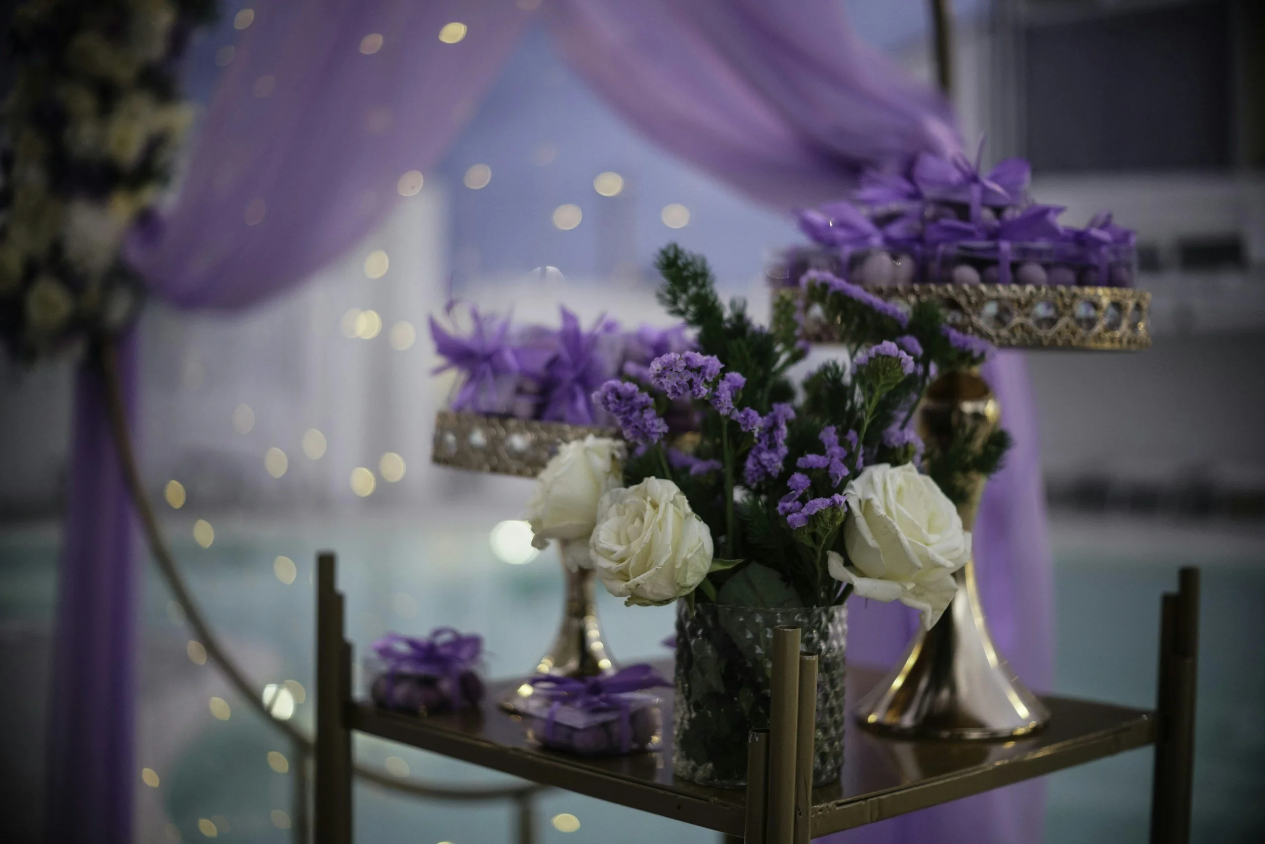 A table decorated with white roses, purple flowers, and purple ribbons, with blurred lights in the background.