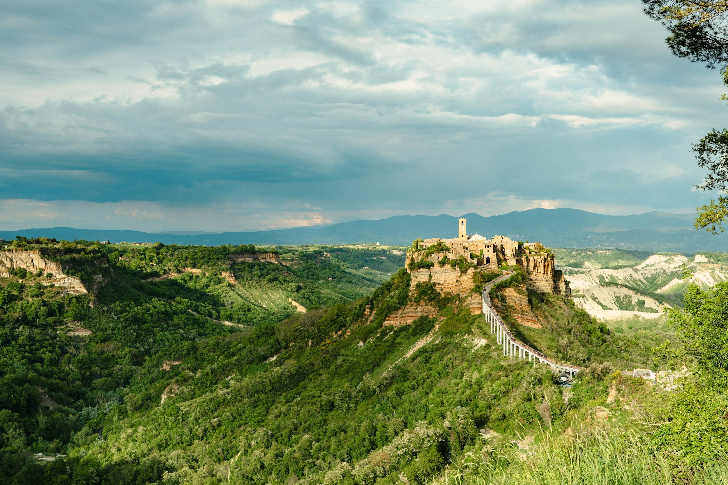 A mountain landscape with a castle-like structure on top of a rocky hill, connected by a bridge, under a cloudy sky, with green vegetation and distant mountains.