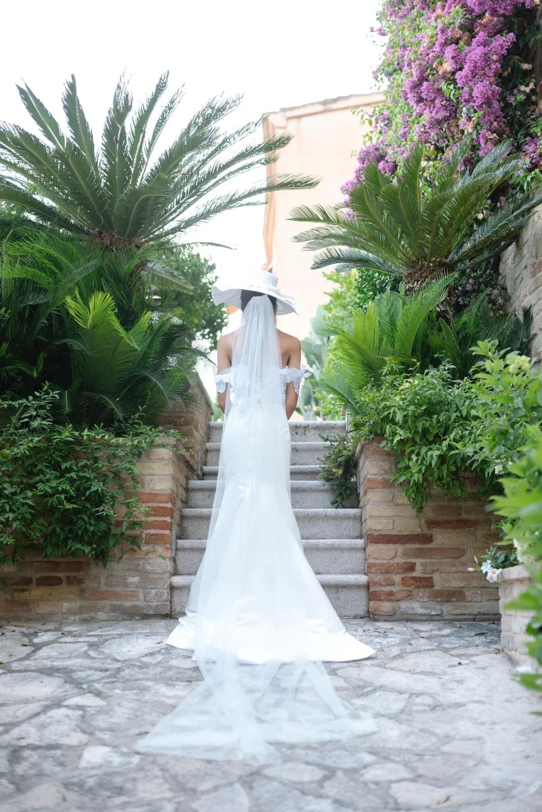 A woman in a white dress, veil, and wide-brimmed hat standing on stone steps surrounded by lush green plants and purple flowers, with a building in the background.