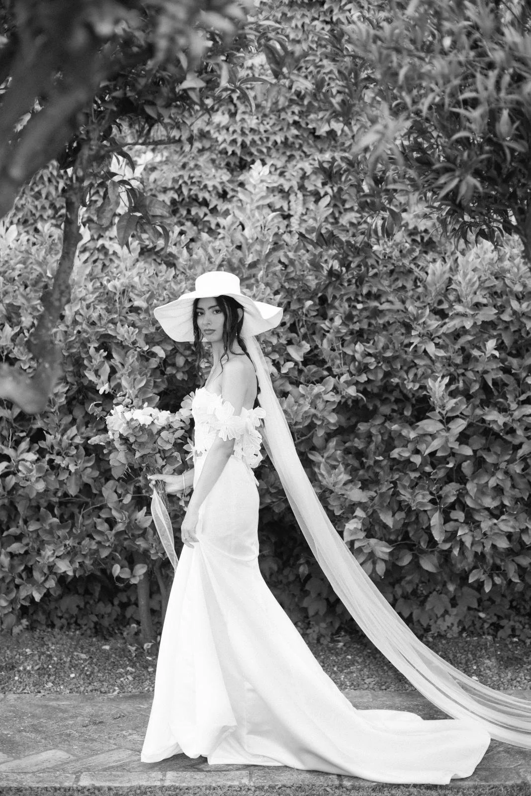 Black and white photo of a woman in a wedding dress and wide-brimmed hat standing outdoors in front of dense foliage.