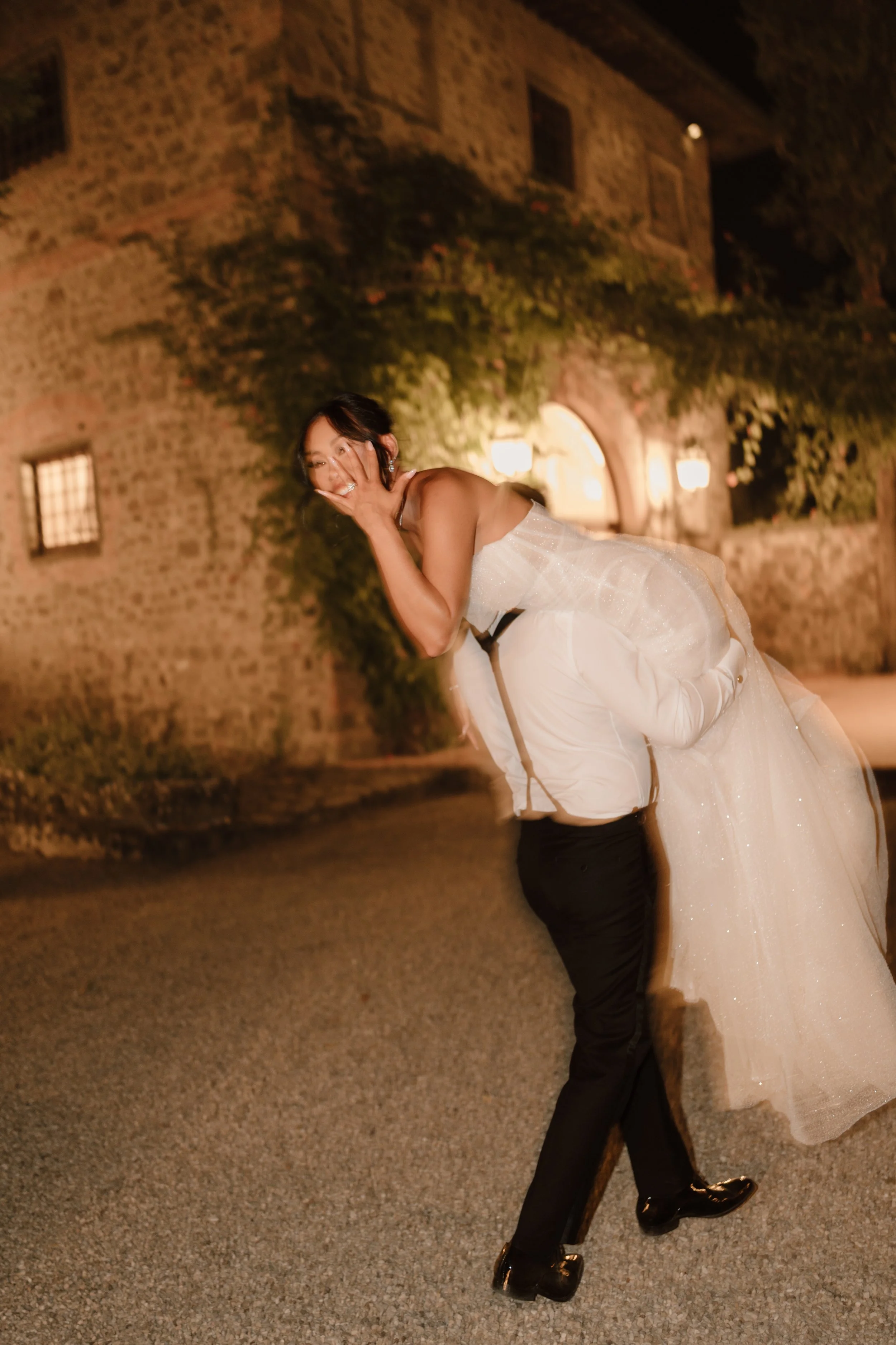 A woman in a wedding dress dancing with a man in a tuxedo outside at night. They are holding each other closely, and the background features a stone building with illuminated windows and greenery.