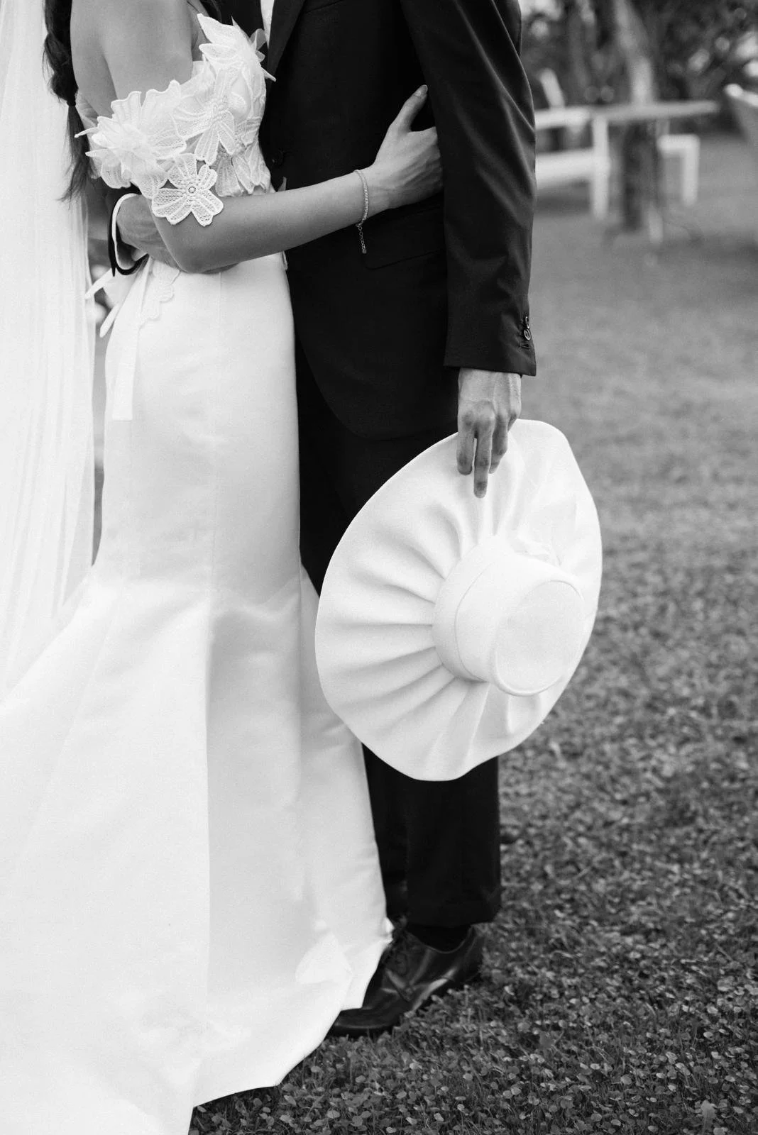 Black-and-white photo of a bride and groom, with the bride wearing a wedding dress with lace details and the groom in a suit holding a wide-brimmed hat.