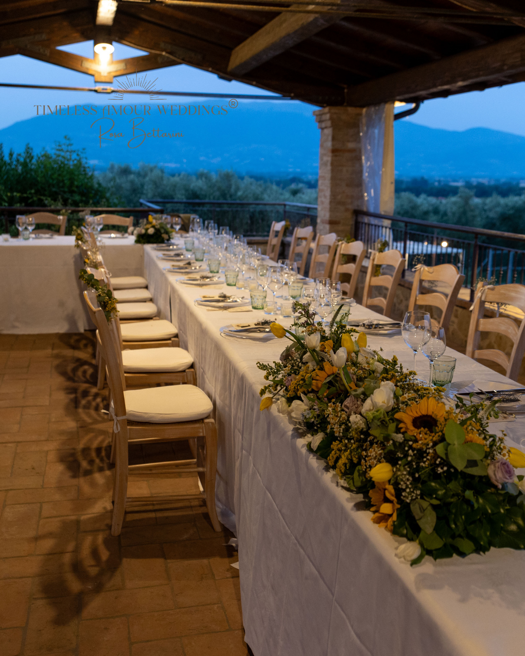 Elegant wedding reception table set with white tablecloth, floral centerpiece, glassware, and cutlery under a wooden pergola with scenic mountain view at dusk.