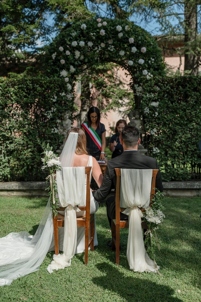 A couple getting married outdoors beneath a flower-covered arch, seated on wooden chairs decorated with white fabric and greenery, with an officiant standing in front of them.