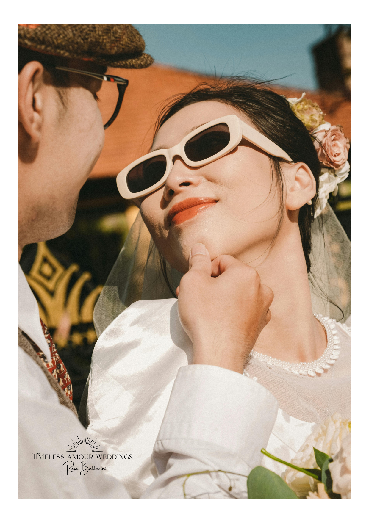 Close-up of a bride and groom, with the groom touching the bride's chin, both wearing sunglasses, during a wedding ceremony outdoors.