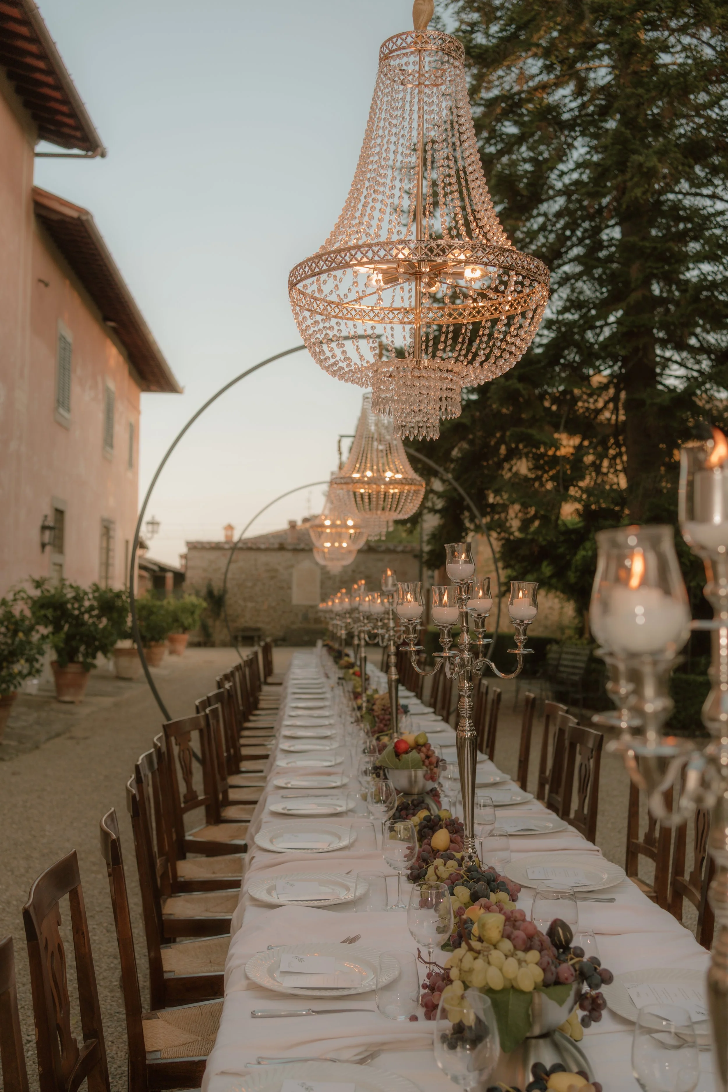 A long outdoor dining table set for a meal, decorated with fruit centerpieces, candles, and elegant glassware. Large crystal chandeliers hang overhead. The scene is in a courtyard with potted plants, surrounding buildings, and trees, under evening sky lighting.