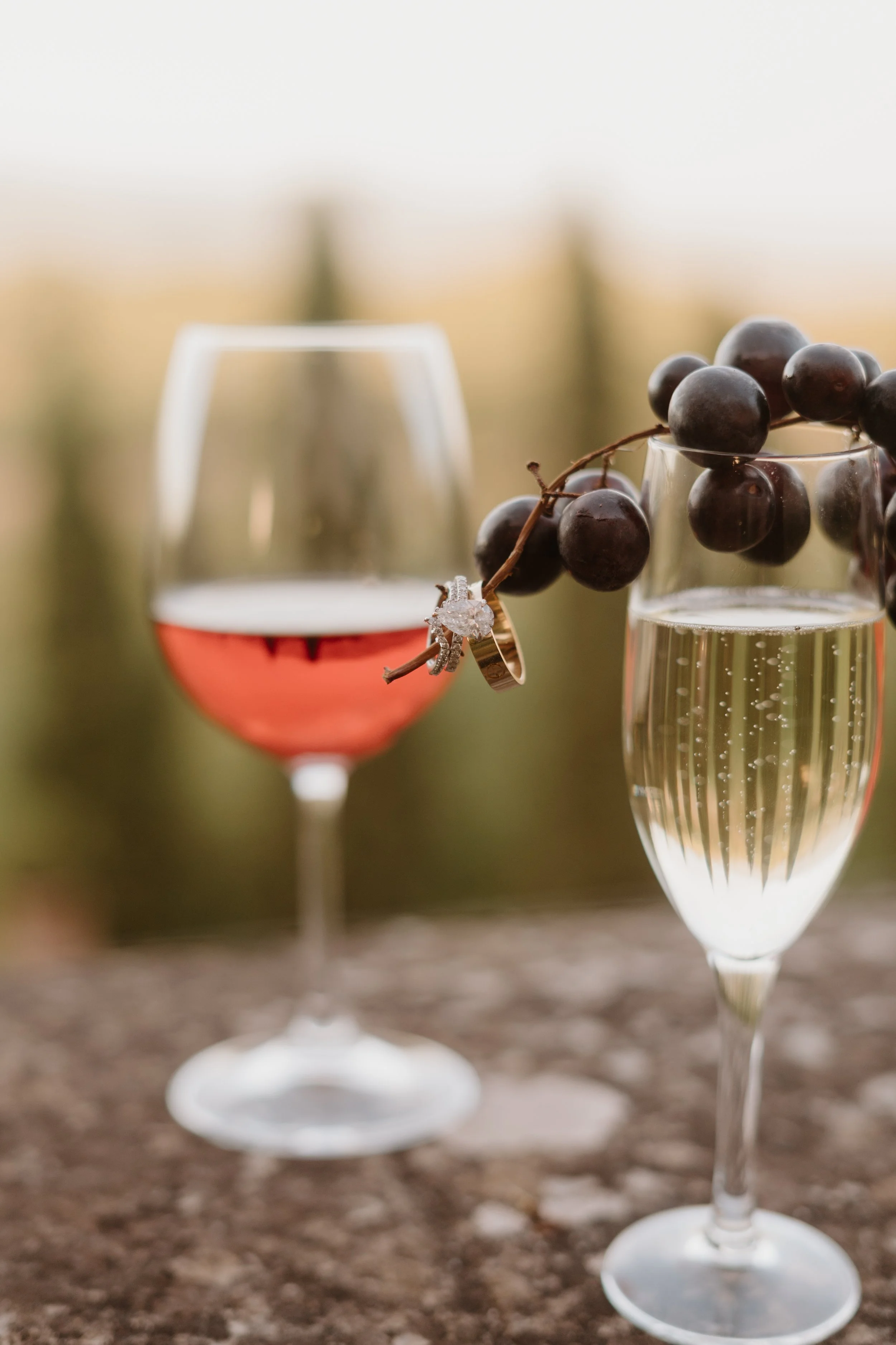 Close-up of a glass of red wine and a glass of champagne with a bunch of black grapes resting on the champagne glass, on a textured surface with a blurred background.