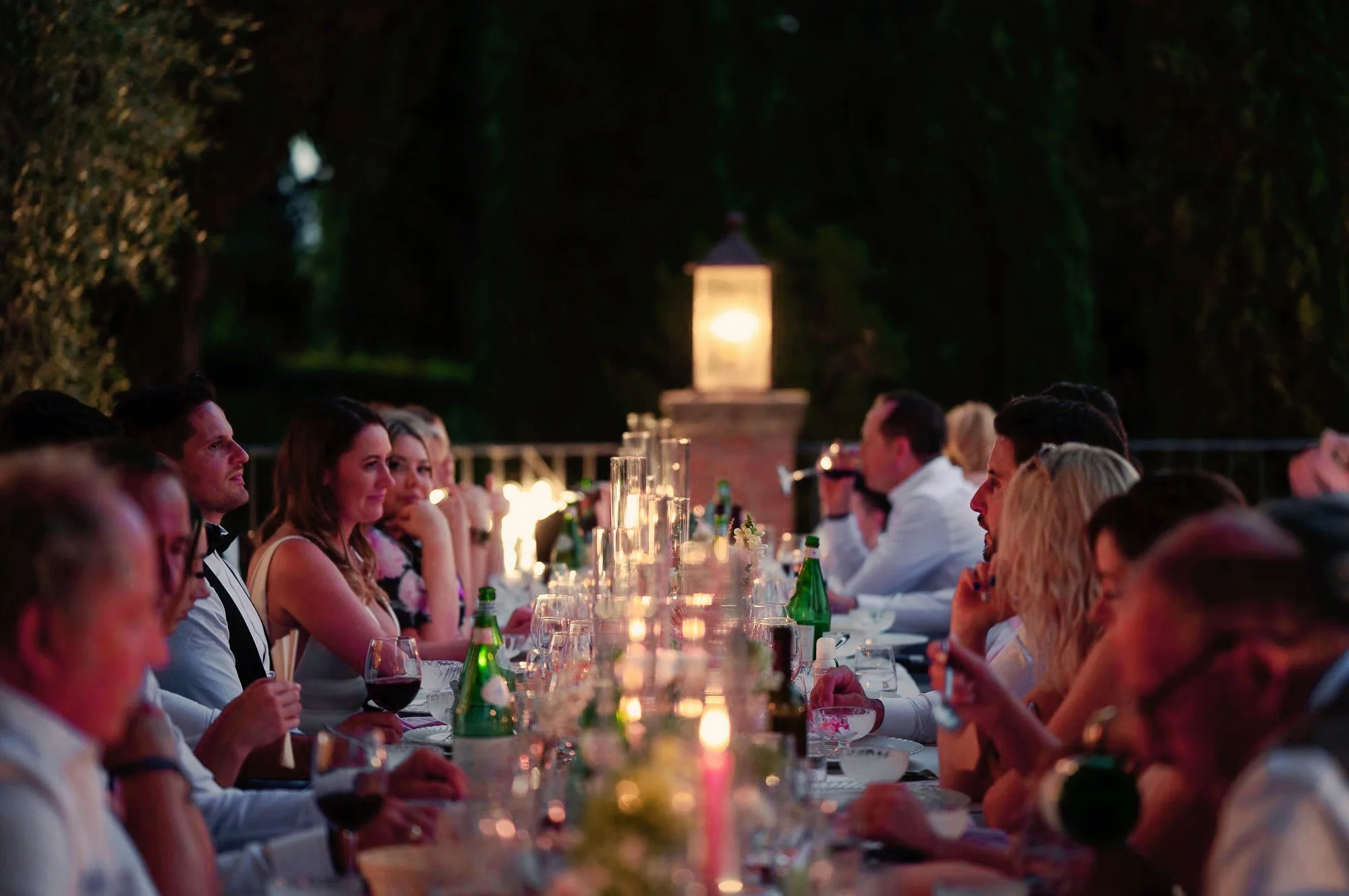 People dining outdoors at night under a canopy of trees, with a lantern in the background.