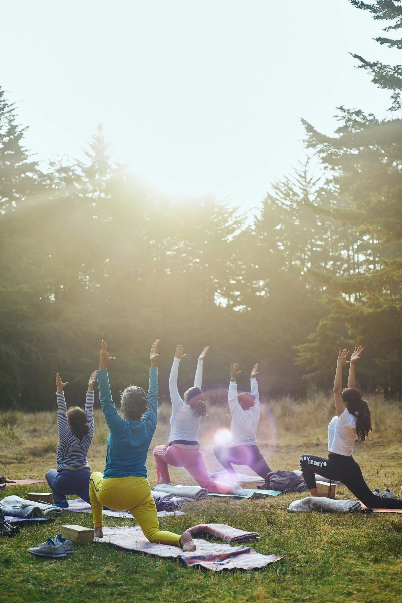 Group of people practicing yoga outdoors in a forest clearing, sun shining through trees, some participants in seated poses, others standing, dressed in comfortable clothing, on yoga mats and blankets.