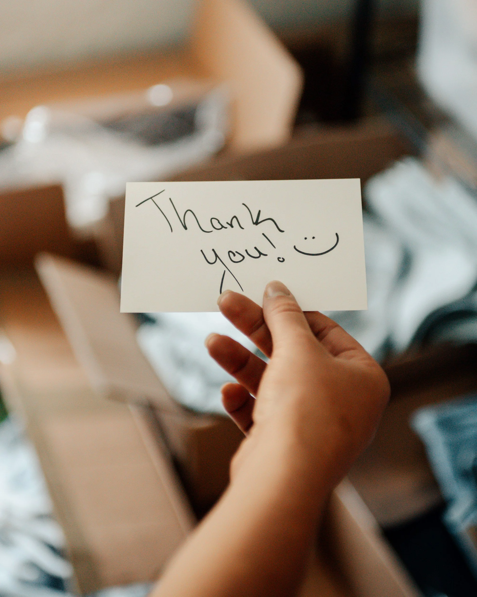A person holding a handwritten note that says 'Thank you!' with a smiley face, in front of a blurred background.