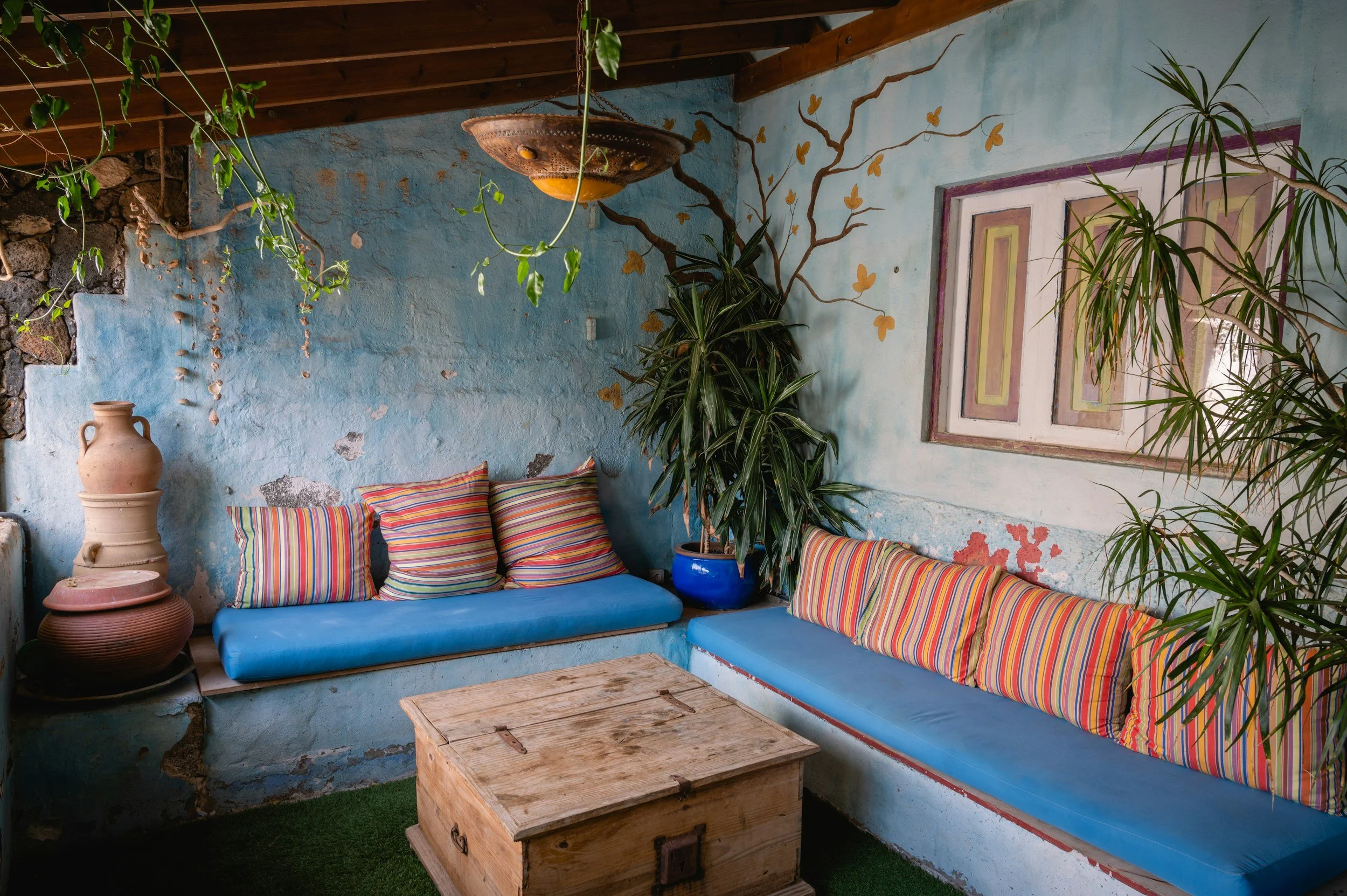 A cozy indoor seating area with a blue cushioned bench decorated with striped pillows, a wooden coffee table, large potted plants, and a wall painted with a mural of a tree with orange leaves on a light blue background.