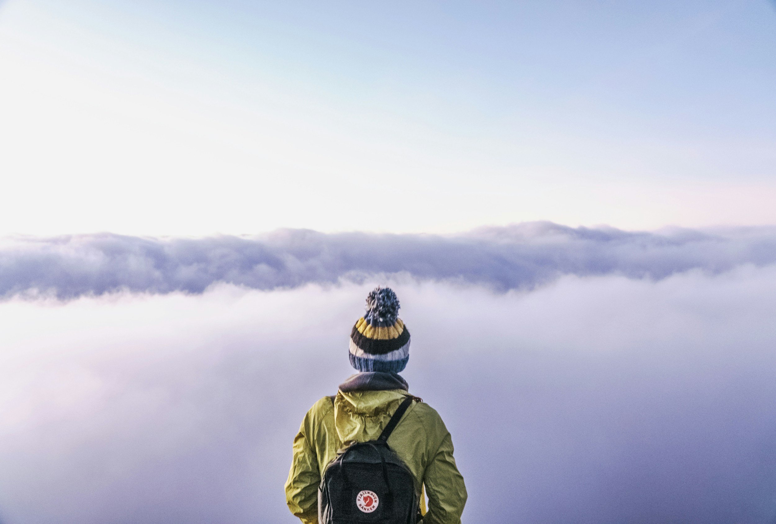 Person standing outdoors with their back to the camera, wearing a yellow jacket, a black backpack, and a knit hat with pom-poms, looking at cloudy sky.