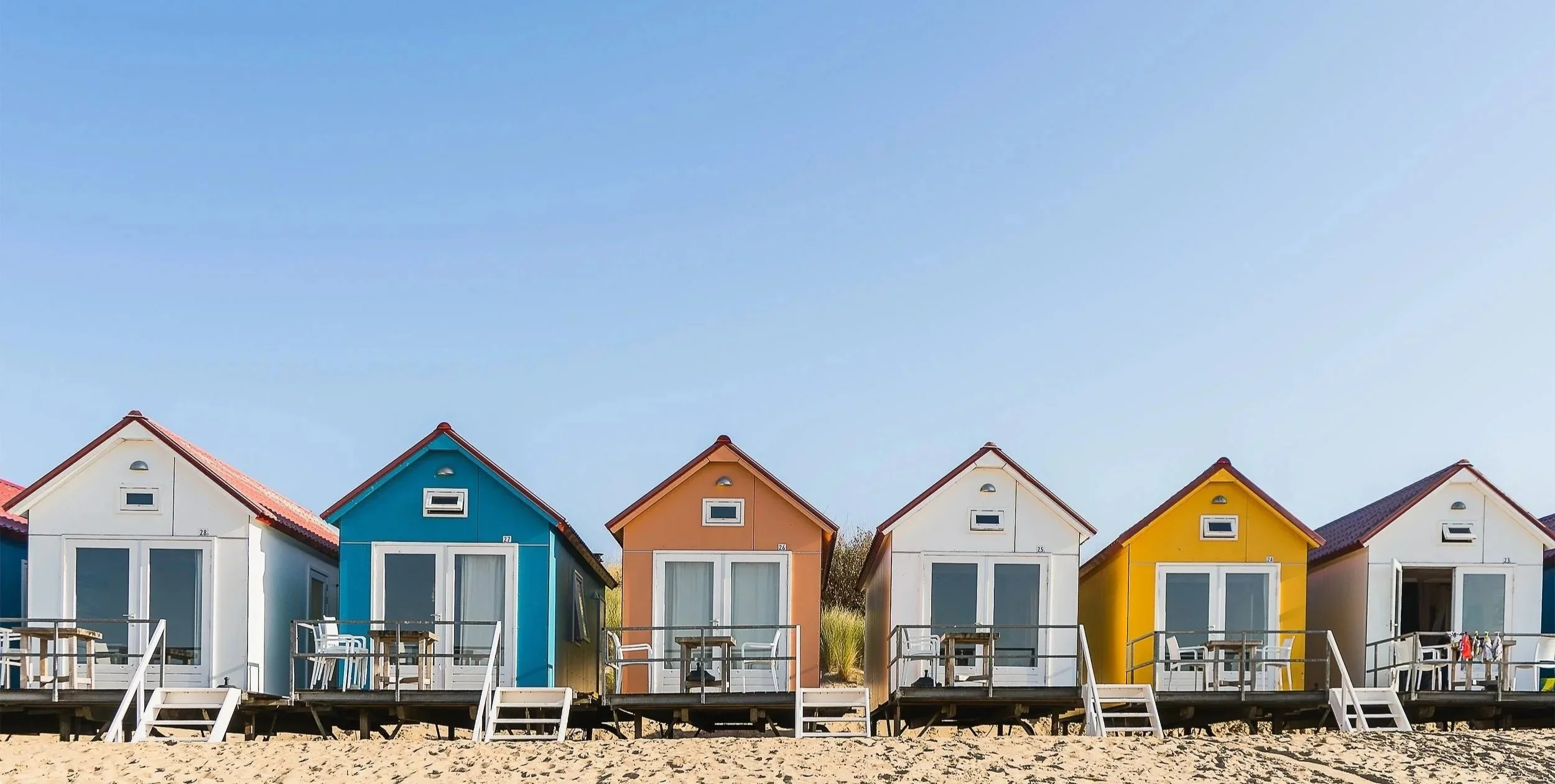 Colorful beach houses in white, blue, orange, yellow, white, and white colors, with stairs leading down to the sandy beach. The houses are on stilts and have small windows, with a clear blue sky in the background.