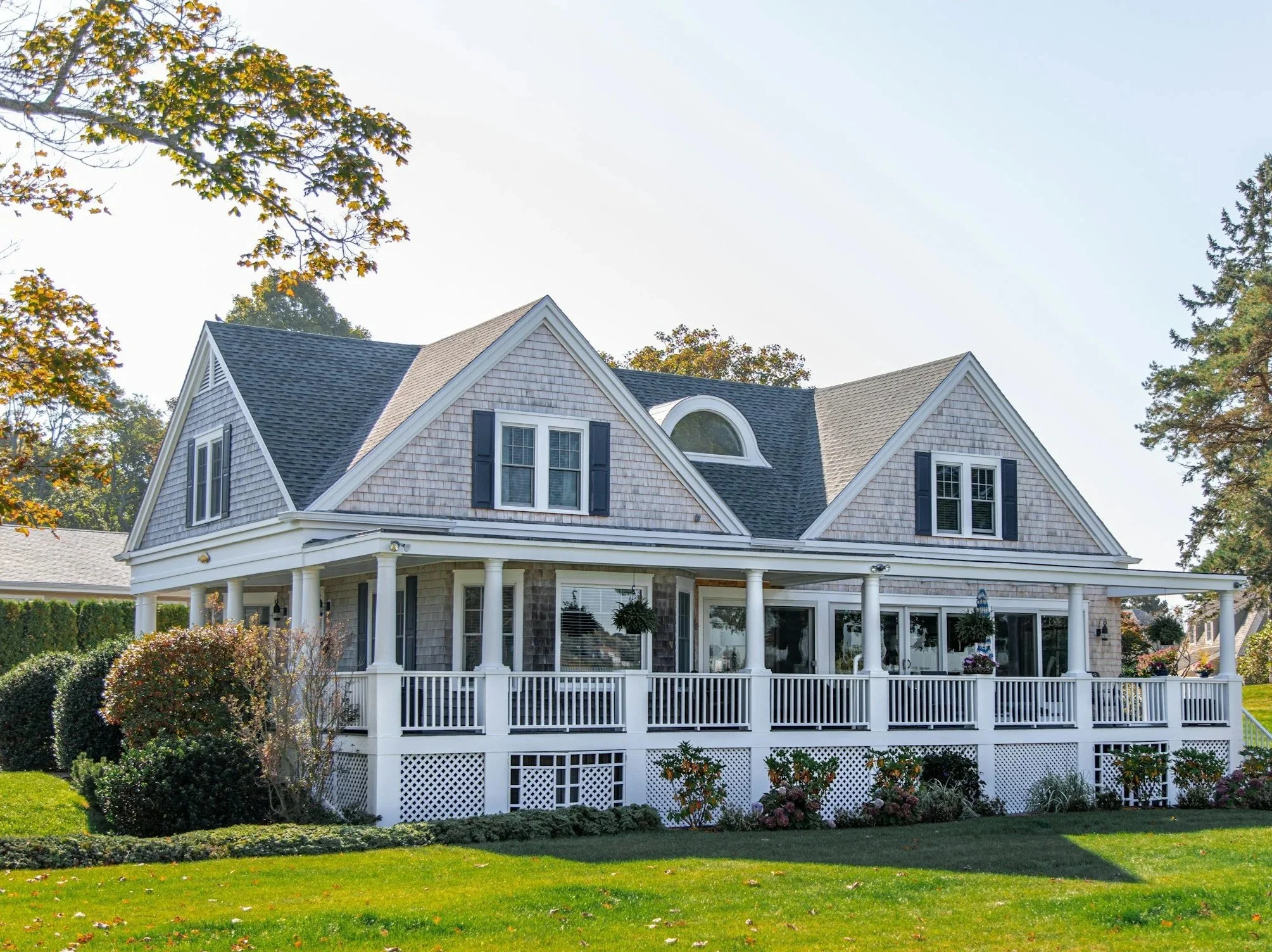 A large two-story house with a wraparound deck, white columns, gray shingles, multiple gabled roofs, and windows with black shutters, surrounded by a well-kept lawn and trees.