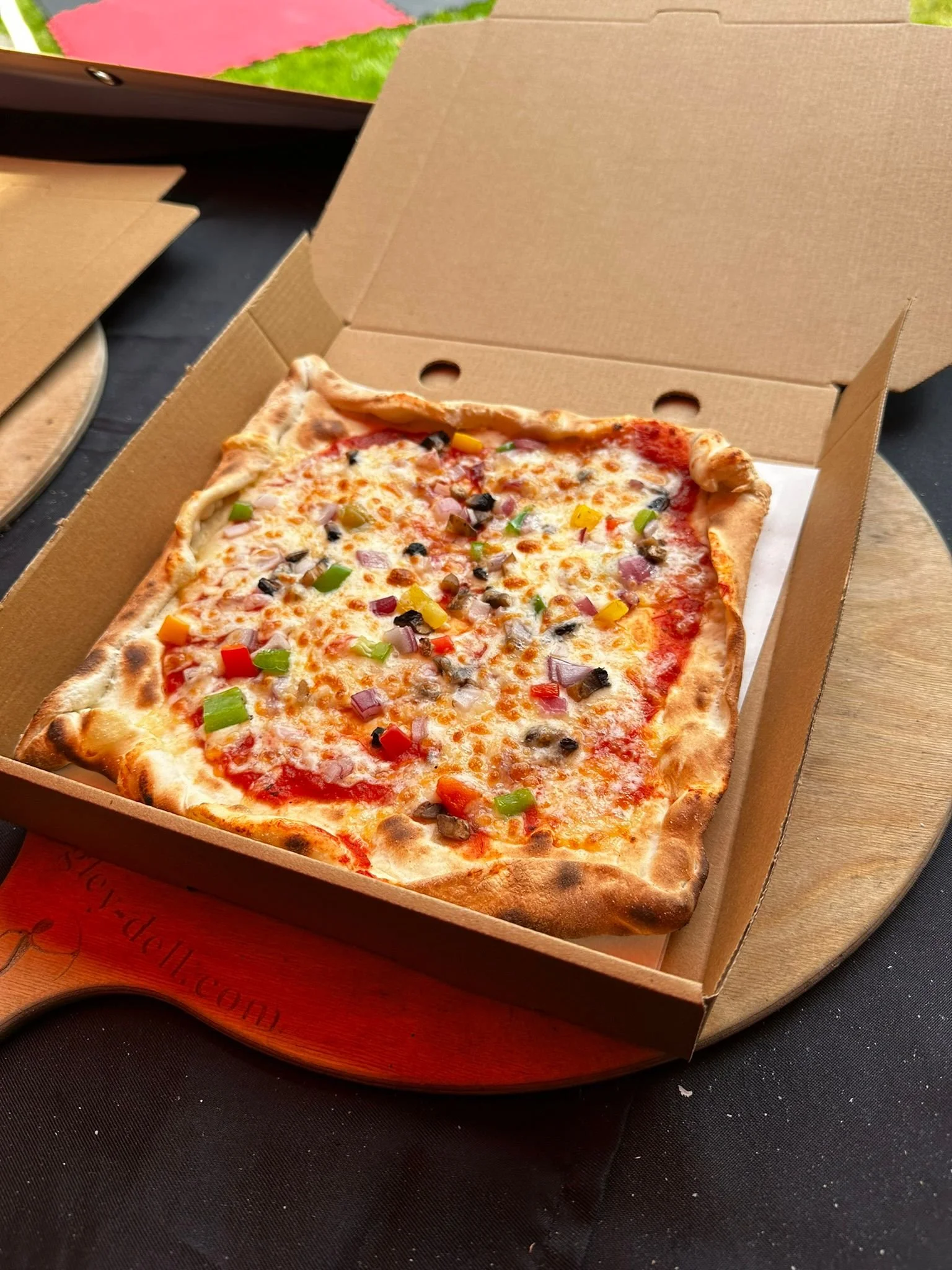 A rectangular pizza with cheese, tomato sauce, and various chopped vegetables in a cardboard pizza box on a round wooden board.