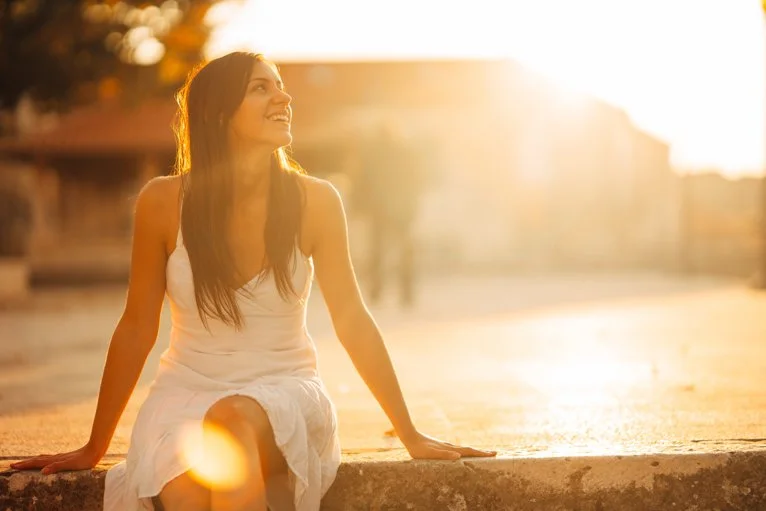 Woman sitting on a stone ledge at sunset, smiling and looking upward.
