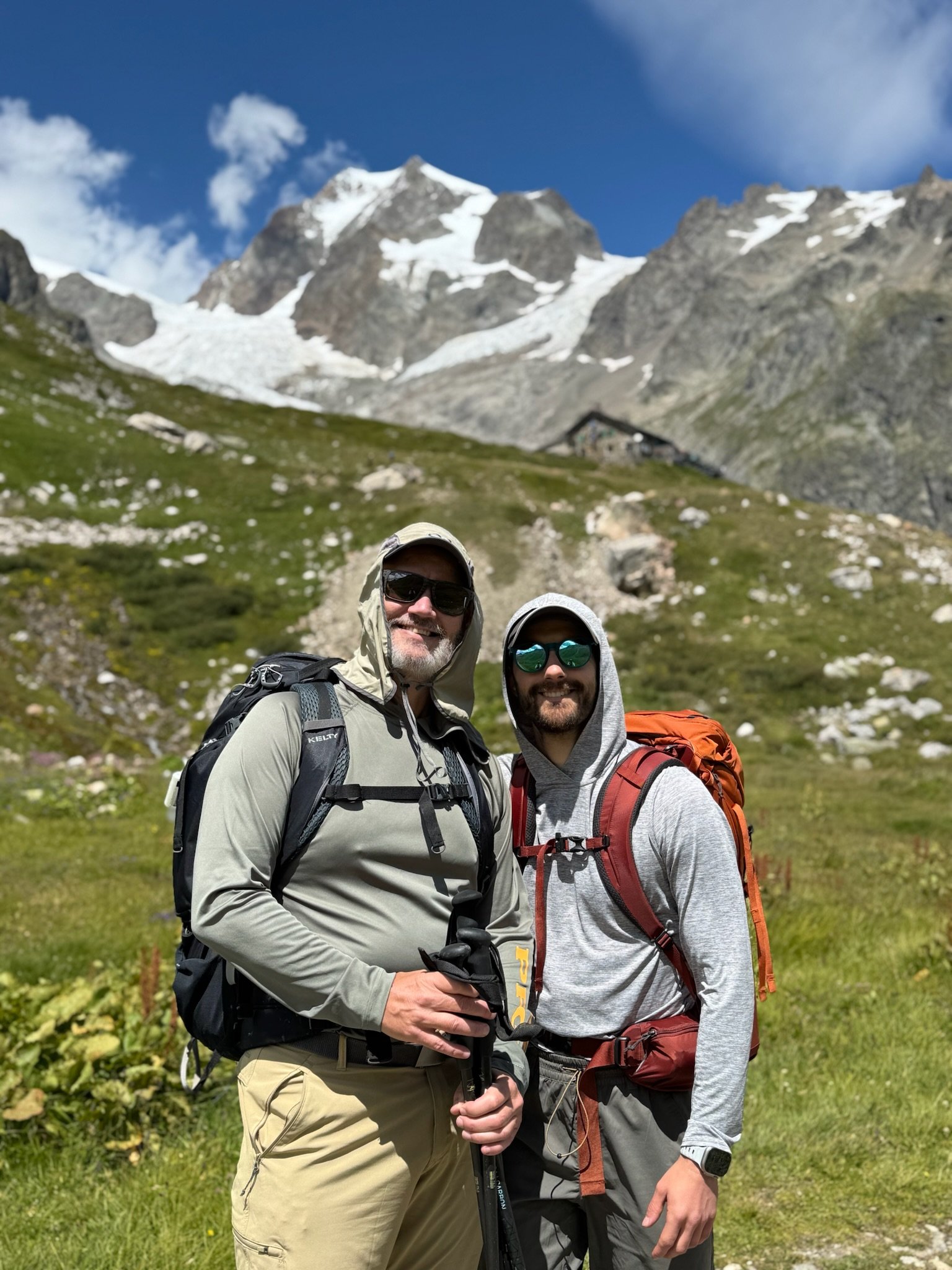 Two men hiking outdoors in a mountainous area with snow-capped peaks in the background, wearing hiking gear and backpacks.