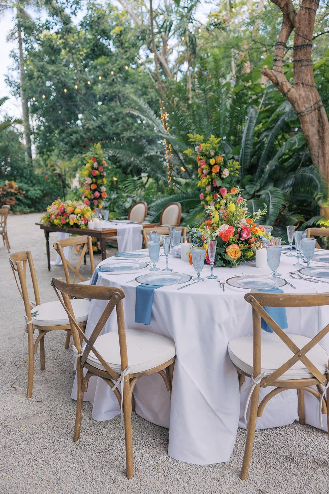 Outdoor wedding reception setup with round tables, floral centerpieces, glassware, and wooden chairs in a garden setting with trees and string lights.