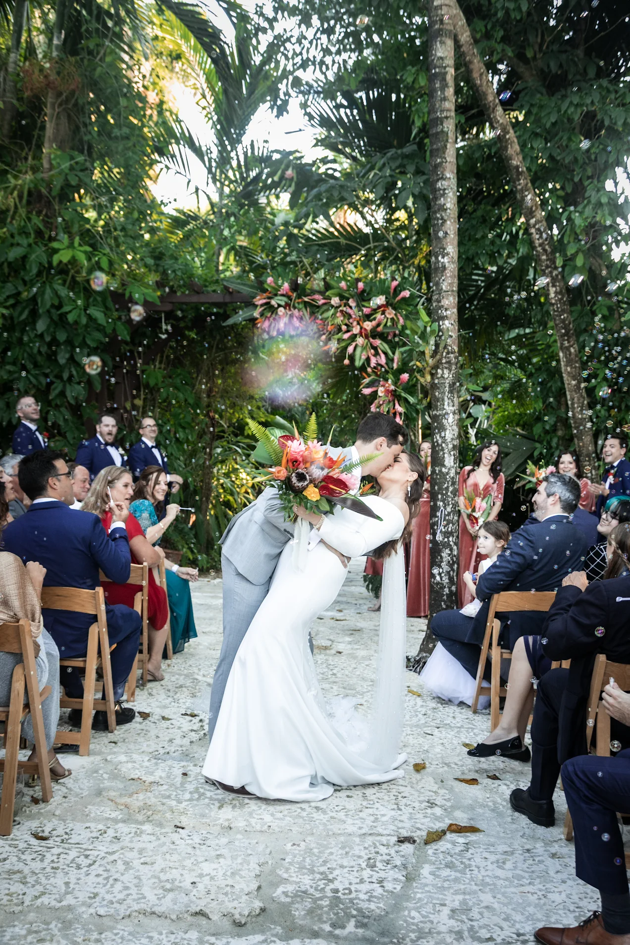 A newlywed couple sharing a kiss during their outdoor wedding ceremony, surrounded by guests and lush tropical greenery.