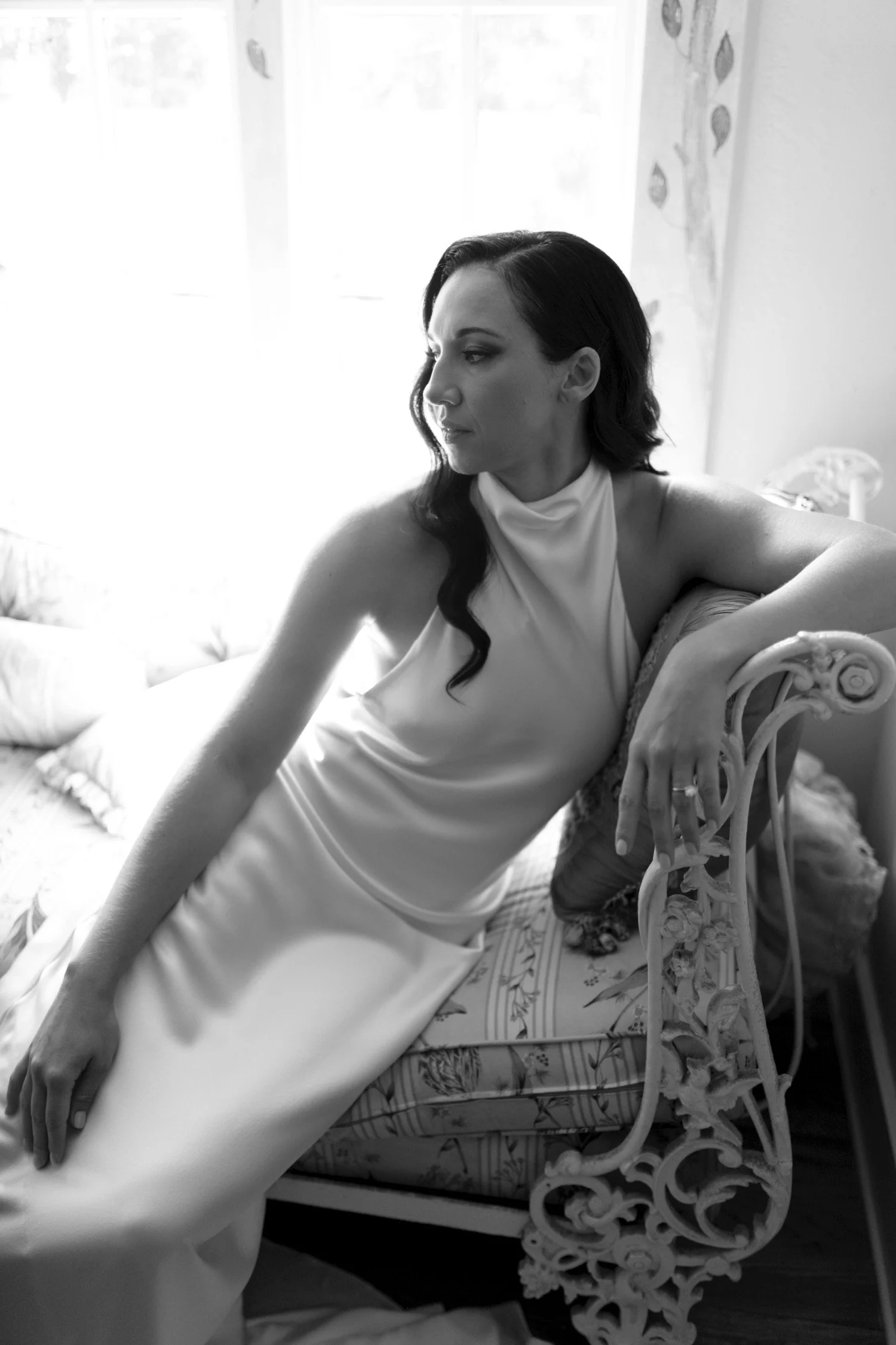 A black and white photo of a woman with dark, wavy hair, wearing a sleeveless, high-neck dress, sitting on a vintage, ornate iron bench with floral cushions, in front of a window with curtains.