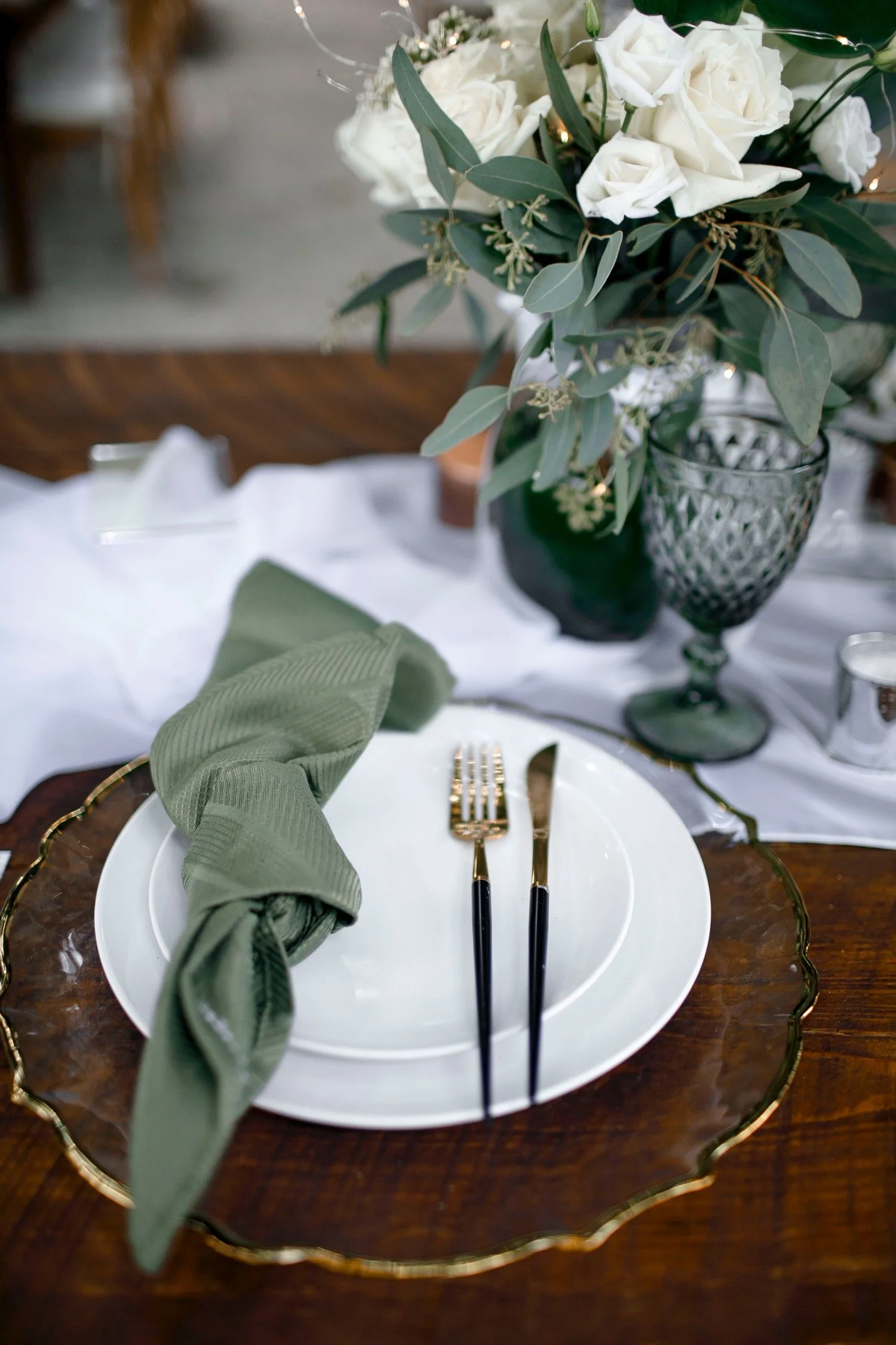 Elegant table setting with green napkin, gold and black cutlery, white plates on a wooden charger plate, and a flower arrangement of white roses and greenery in a dark green vase.