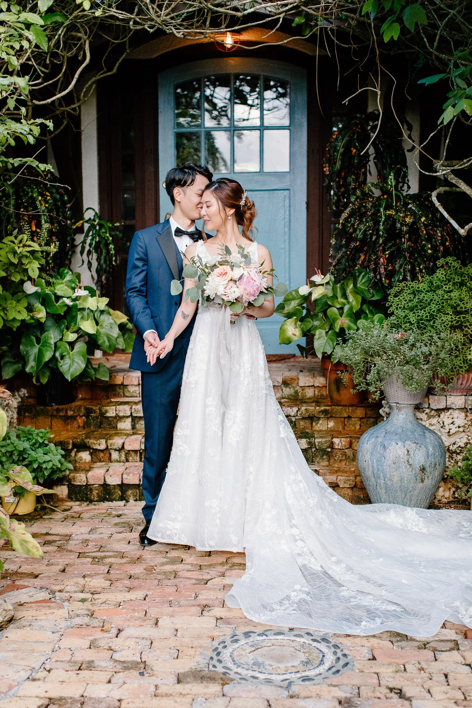 A newlywed couple stands on a brick pathway in front of a blue door, surrounded by lush greenery. The groom in a blue tuxedo gently kisses the bride on her forehead, who wears a white lace wedding gown with a long train and holds a bouquet of flowers