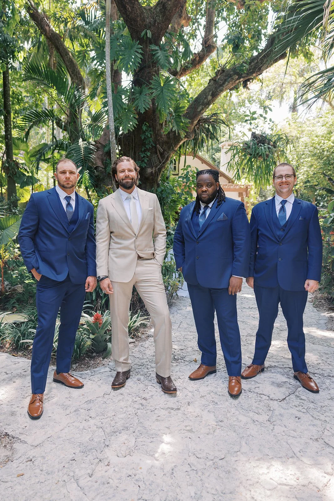 Four men in suits standing under a large tree in a lush garden