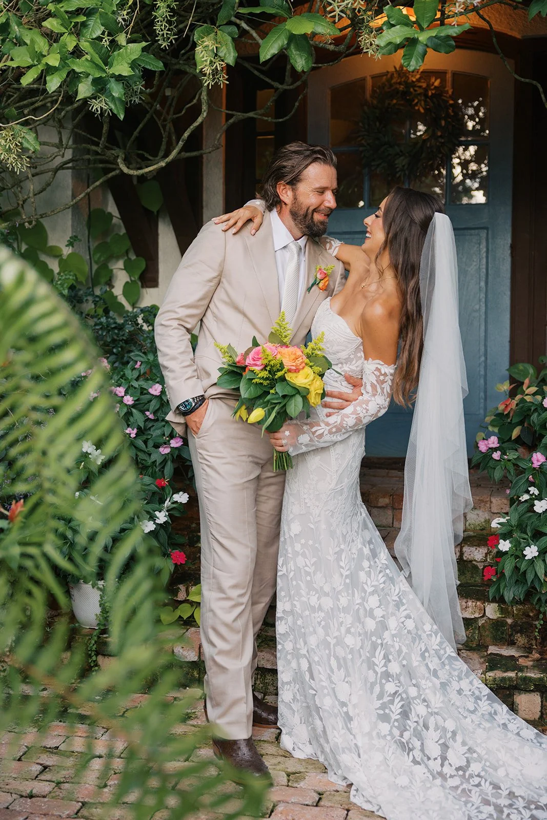A bride and groom share a joyful moment on their wedding day, standing on brick steps surrounded by lush greenery and colorful flowers, with the bride holding a bouquet of yellow and pink roses.