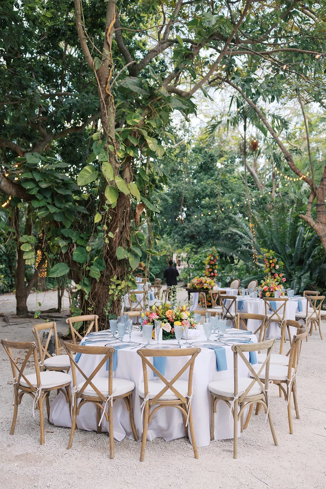 Outdoor garden wedding or event setup with round tables decorated with floral centerpieces, white tablecloths, and blue napkins, surrounded by wooden chairs with white cushions, under large trees with string lights.