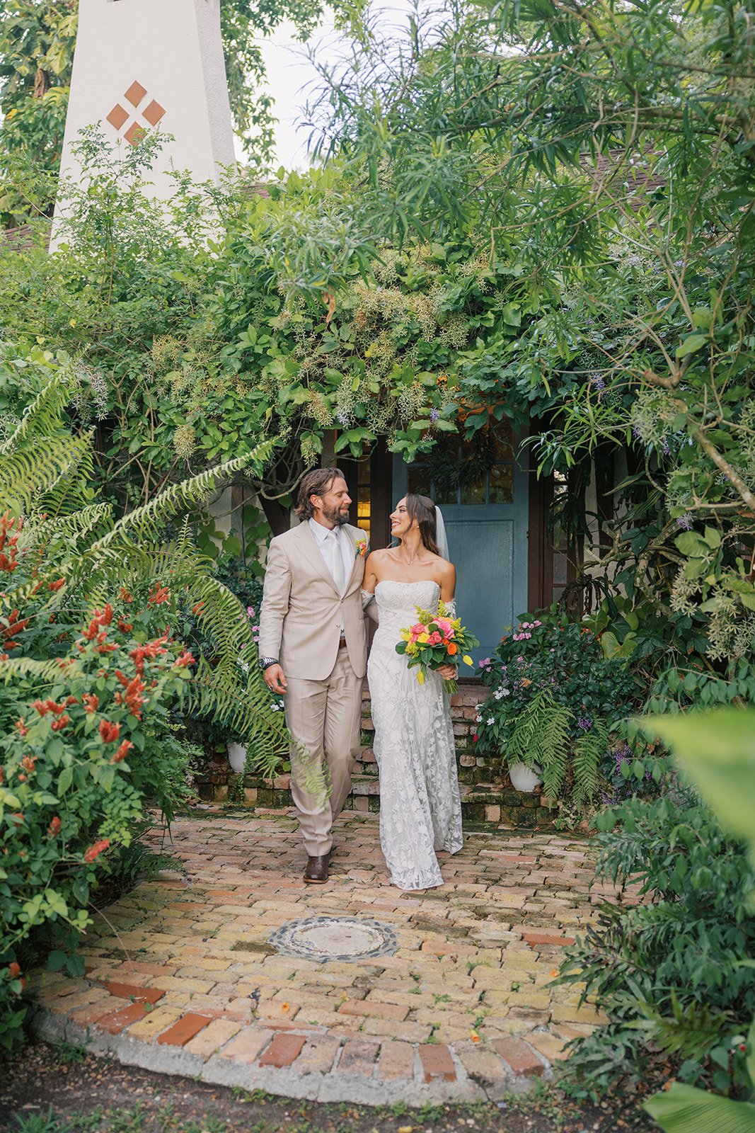A bride and groom walking arm in arm in a lush garden, surrounded by green foliage and colorful flowers, with a blue door in the background.